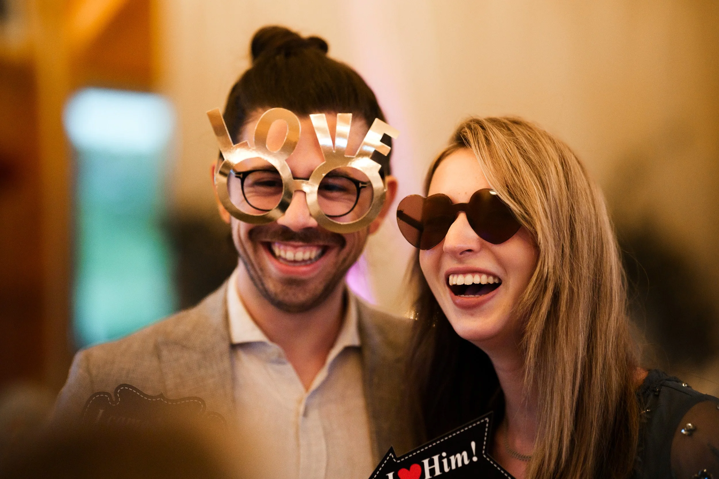Ein lachendes Paar bei einer Party, beide tragen sunglasses, der Mann hat eine goldene Brille mit 'LOVE' und der Frau eine Herz-Sonnenbrille.