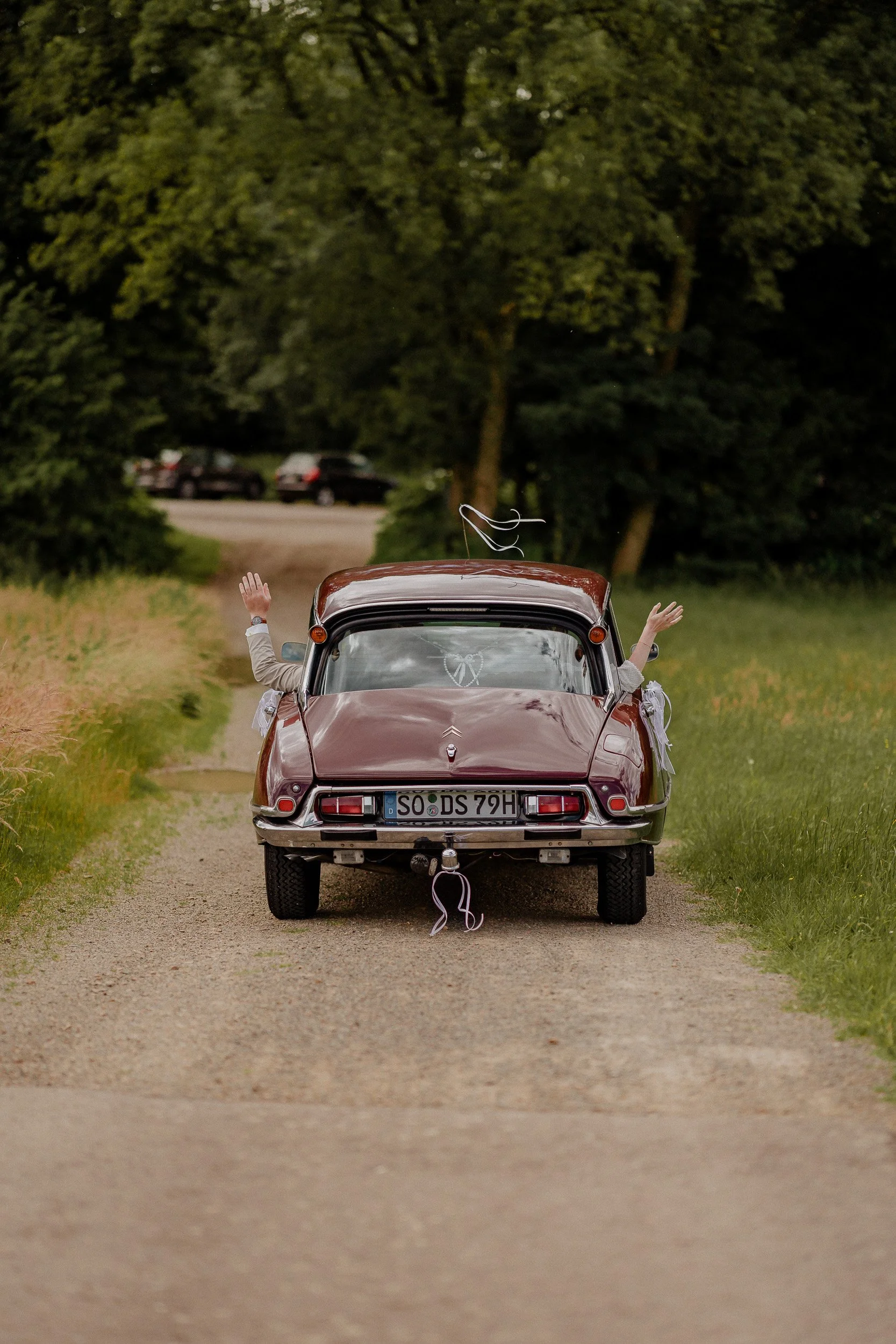 Ein rotes Auto auf einem Feldweg, mit einer Person, die vorne aus dem Fahrzeug mit hochgehobenen Armen heraushockt, vermutlich bei einer Hochzeit.