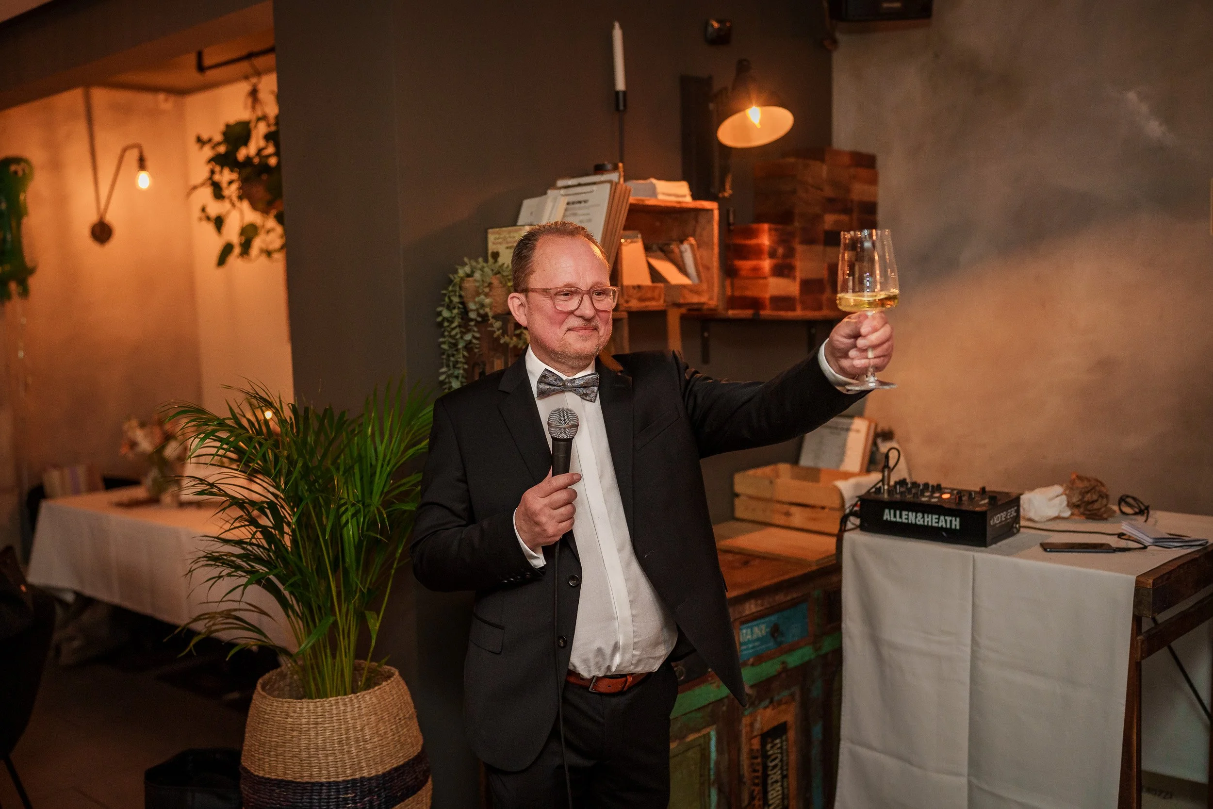 Ein Mann in einem schwarzen Anzug mit Fliege hält ein Mikrofon in einer Hand und ein Wein Glas in der anderen, während er eine Rede hält.
