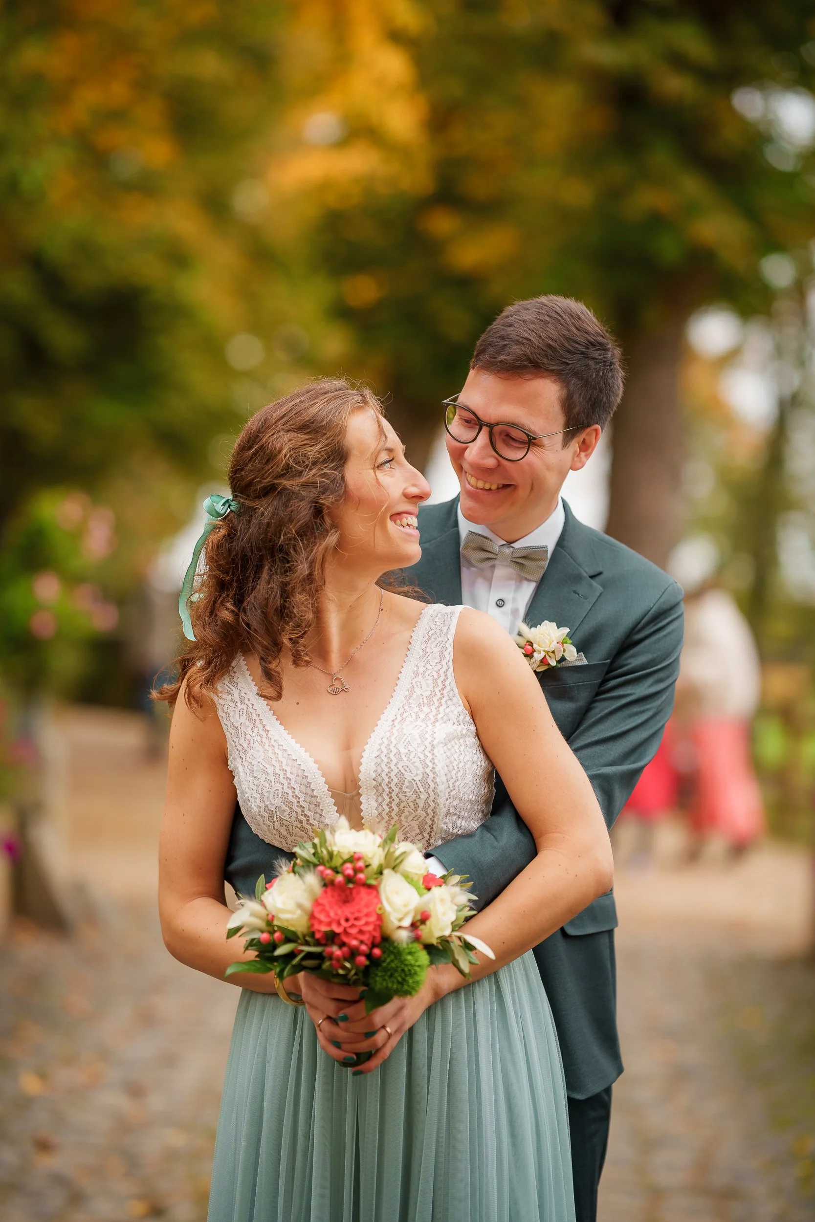 Ein glückliches Brautpaar bei einer Hochzeit im Freien, die Braut hält einen Blumenstrauß, beide lachen sich an, im Hintergrund sind Bäume mit herbstlichen Blättern | Fabian Dick Hochzeitsfotograf | Burg Adendorf
