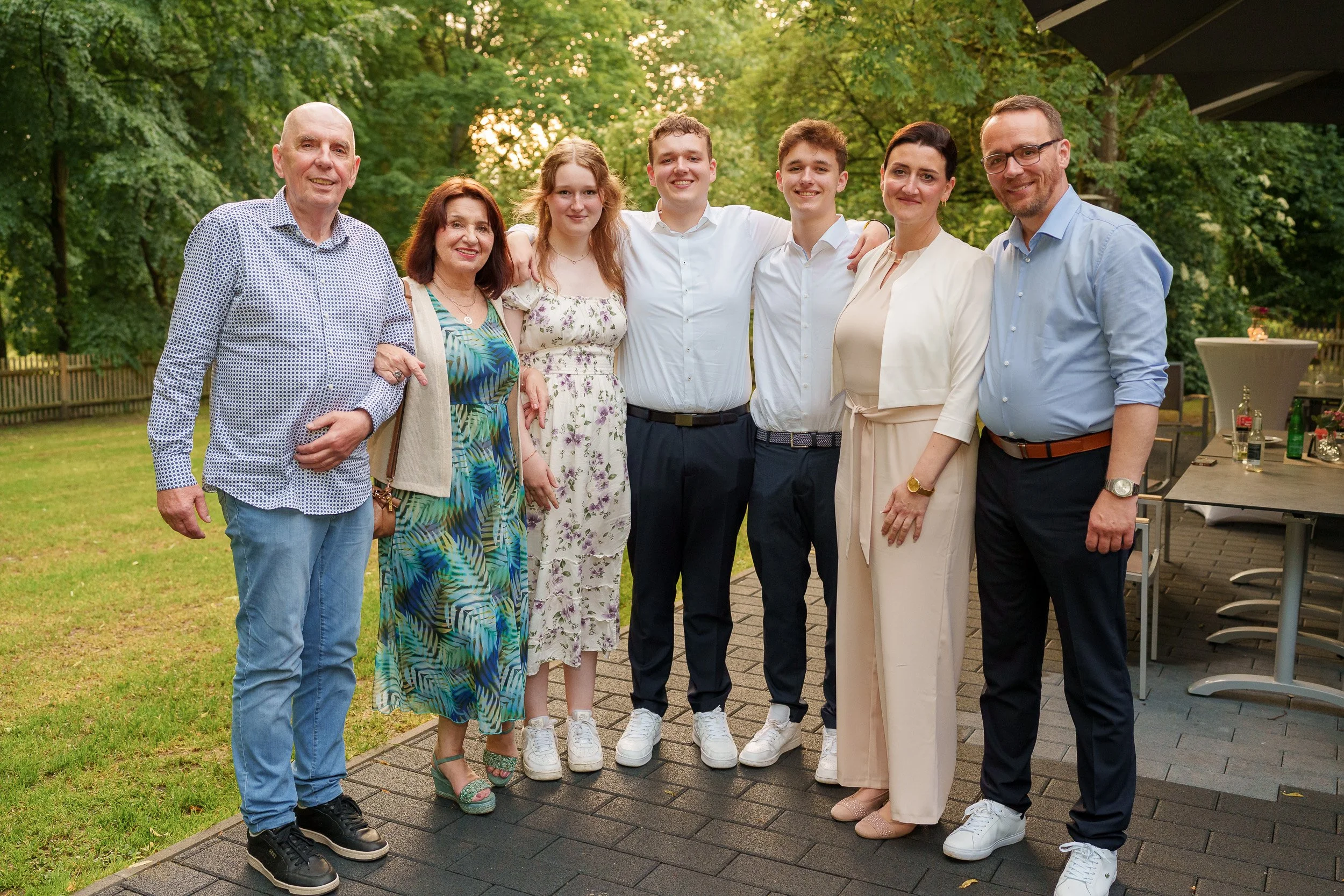 Eine Gruppe von acht Menschen steht draußen auf einem Rasen mit Bäumen im Hintergrund, bei Sonnenuntergang, sie lächeln und haben Arme um die Schultern, feierliche Kleidung, vermutlich Familien- oder Freundestreffen.