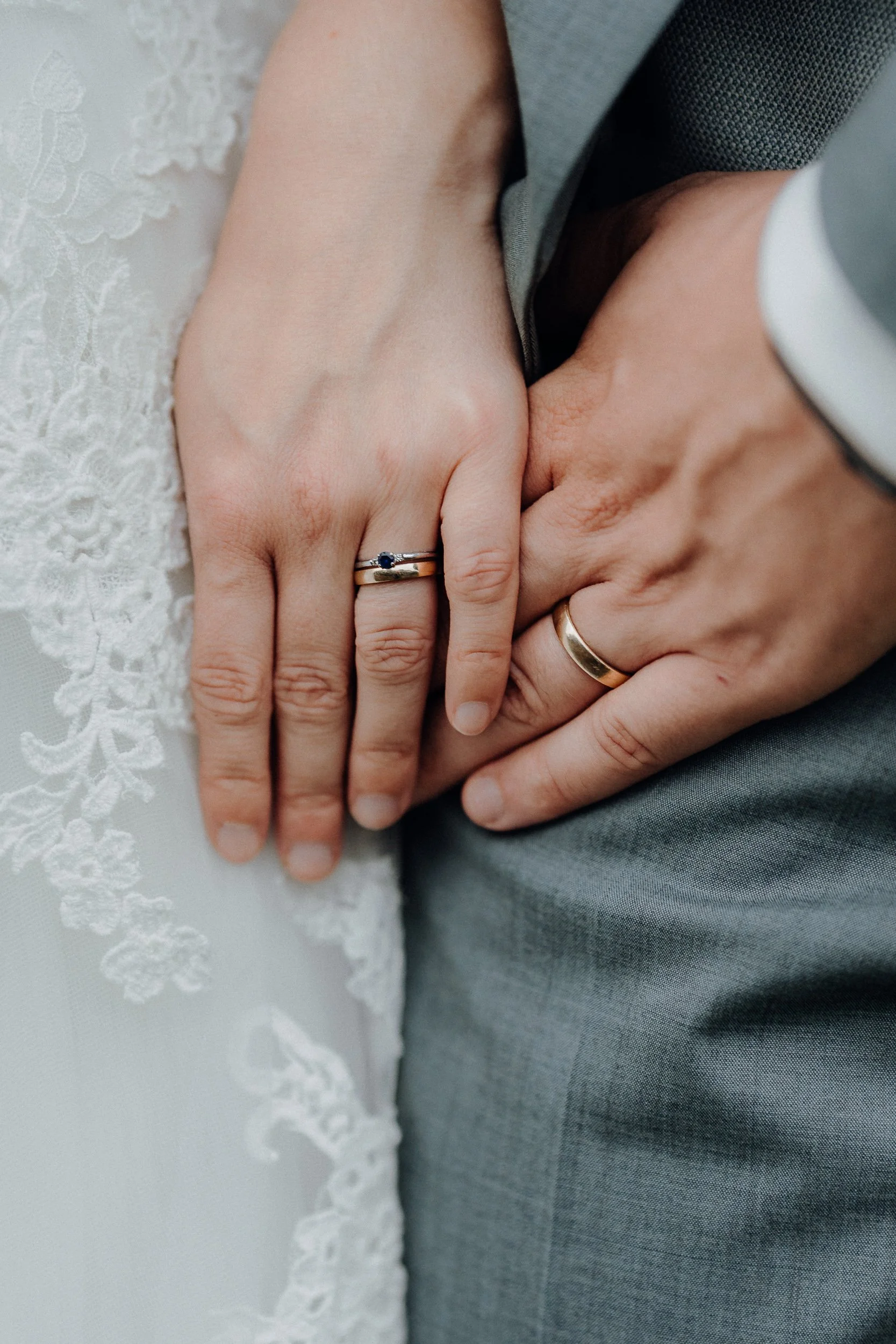 Hände eines verheirateten Paares mit Eheringen, die auf einem Hochzeitskleid und einem Anzug ruhen.