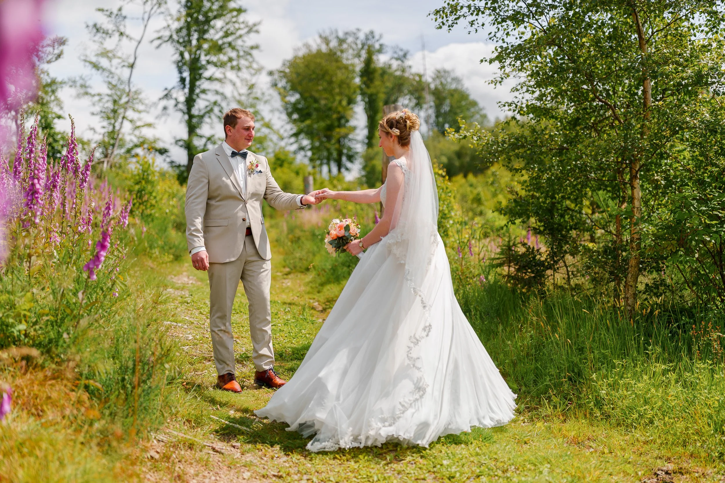 Ein Brautpaar steht in einem Grünbereich, die Braut trägt ein weißes Hochzeitskleid und hält einen Blumenstrauß, der Bräutigam trägt einen beigen Anzug und eine Fliege. Sie scheinen im Sonnenlicht zu stehen, umgeben von Bäumen und Blumen.