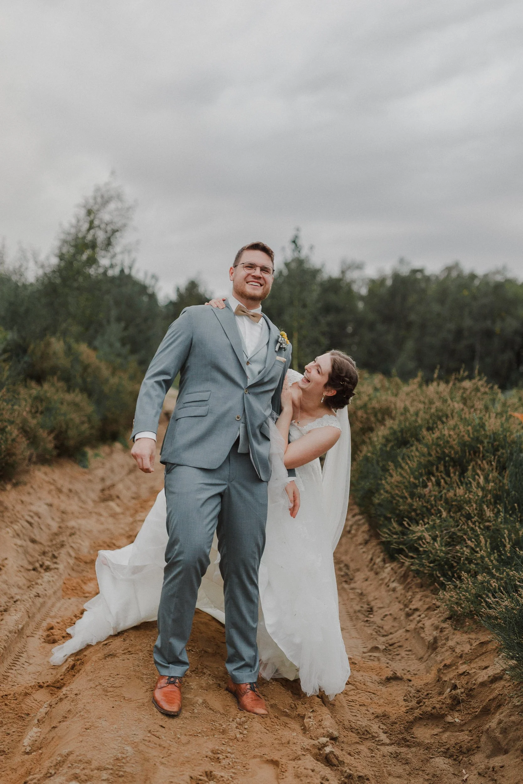 Ein lachendes Brautpaar steht auf einem Sandweg in einer natürlichen, grünen Umgebung bei bewölktem Himmel. Der Bräutigam trägt einen grauen Anzug und die Braut trägt ein weißes Hochzeitskleid.