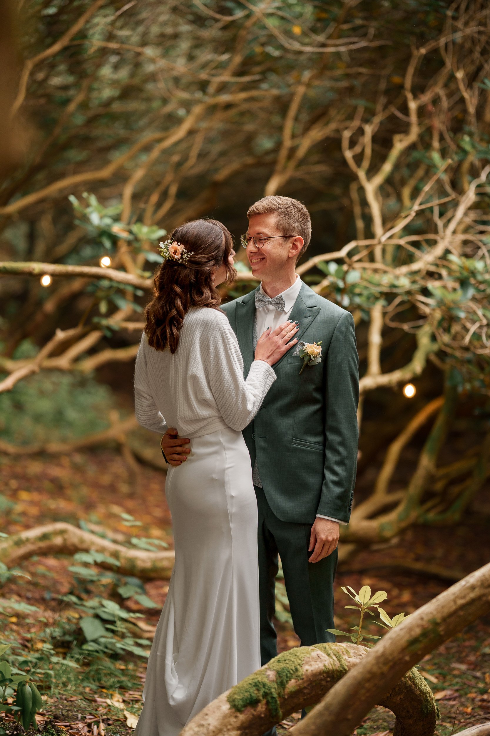 Ein Brautpaar steht in einem Wald, lächelt sich an und hält sich an den Händen. Die Braut trägt ein langes weißes Kleid und hat einen Blumenkranz im Haar, der Bräutigam trägt einen grünen Anzug mit Fliege. Im Hintergrund sind Bäume und kleine Lichter