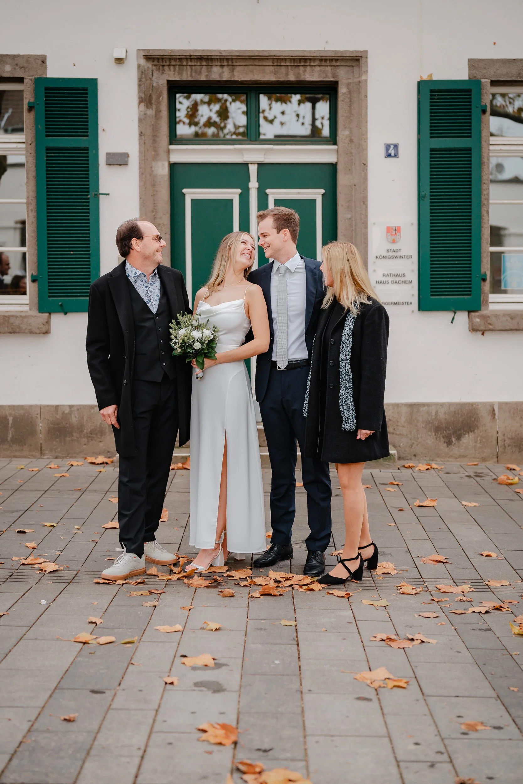 Vier Menschen stehen auf einem Gehweg vor einem Gebäude mit grünen Fensterläden. Es ist Herbst, Blätter liegen auf dem Boden. Zwei Frauen und zwei Männer, die an einer Hochzeit beteiligt zu sein scheinen, lachen und schauen sich an. Die Frau in der M