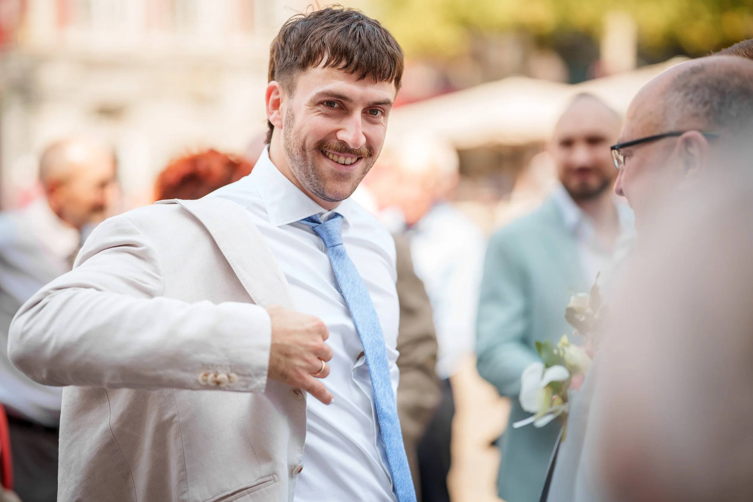 Ein Mann in einem weißen Hemd und blauer Krawatte lächelt in die Kamera, während er eine beige Jacke trägt, auf einem gesellschaftlichen Event im Freien im Hintergrund sind andere Menschen mit hellen Hemden und Anzügen.