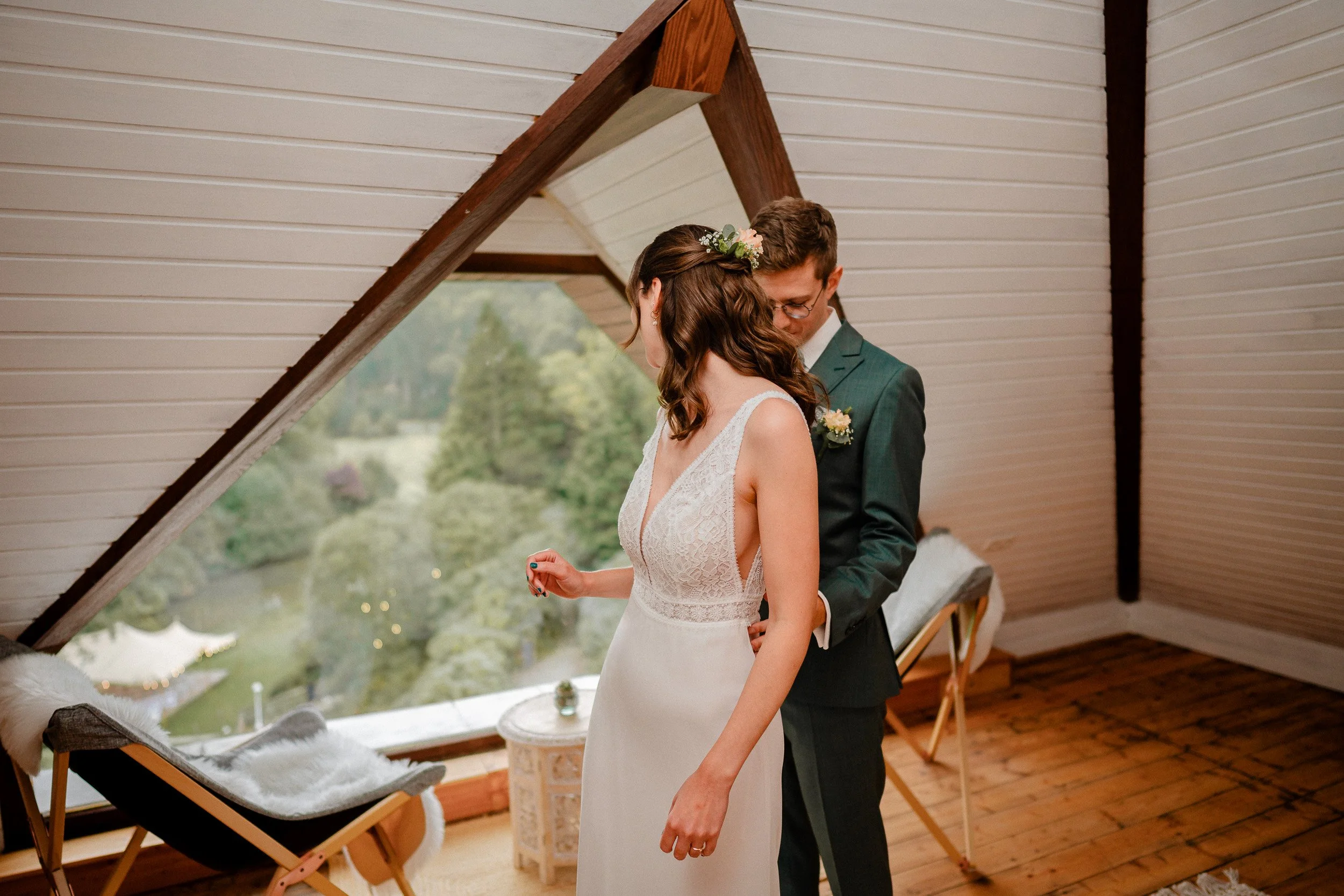 Ein Brautpaar steht in einem Dachzimmer vor einem großen Fenster mit Blick auf einen Garten. Es ist ein intimer Moment während einer Hochzeit, umgeben von Holzdecken und stilvoller Einrichtung.