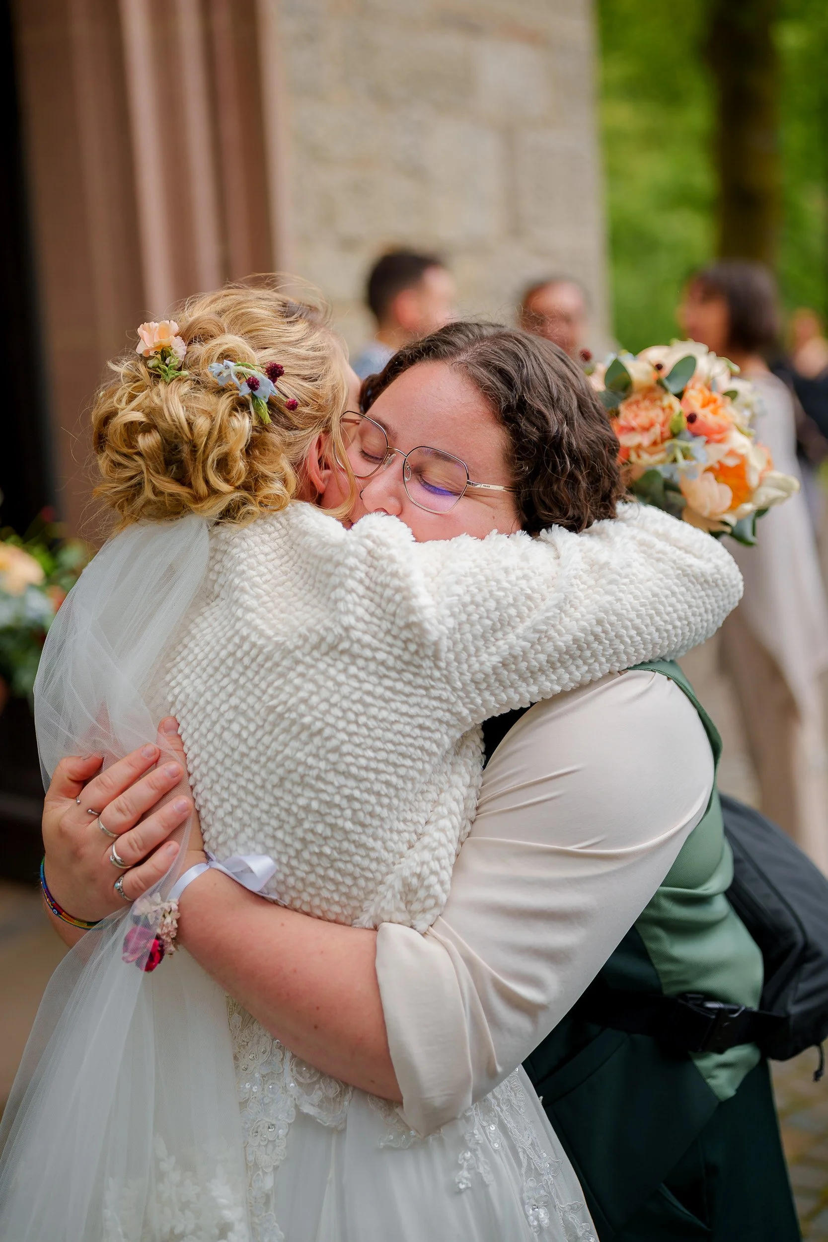 Zwei Frauen umarmen sich bei einer Hochzeit, eine trägt ein weißes Brautkleid mit Schleier, die andere hat lockiges Haar, trägt eine grüne Weste und hält einen Blumenstrauß.