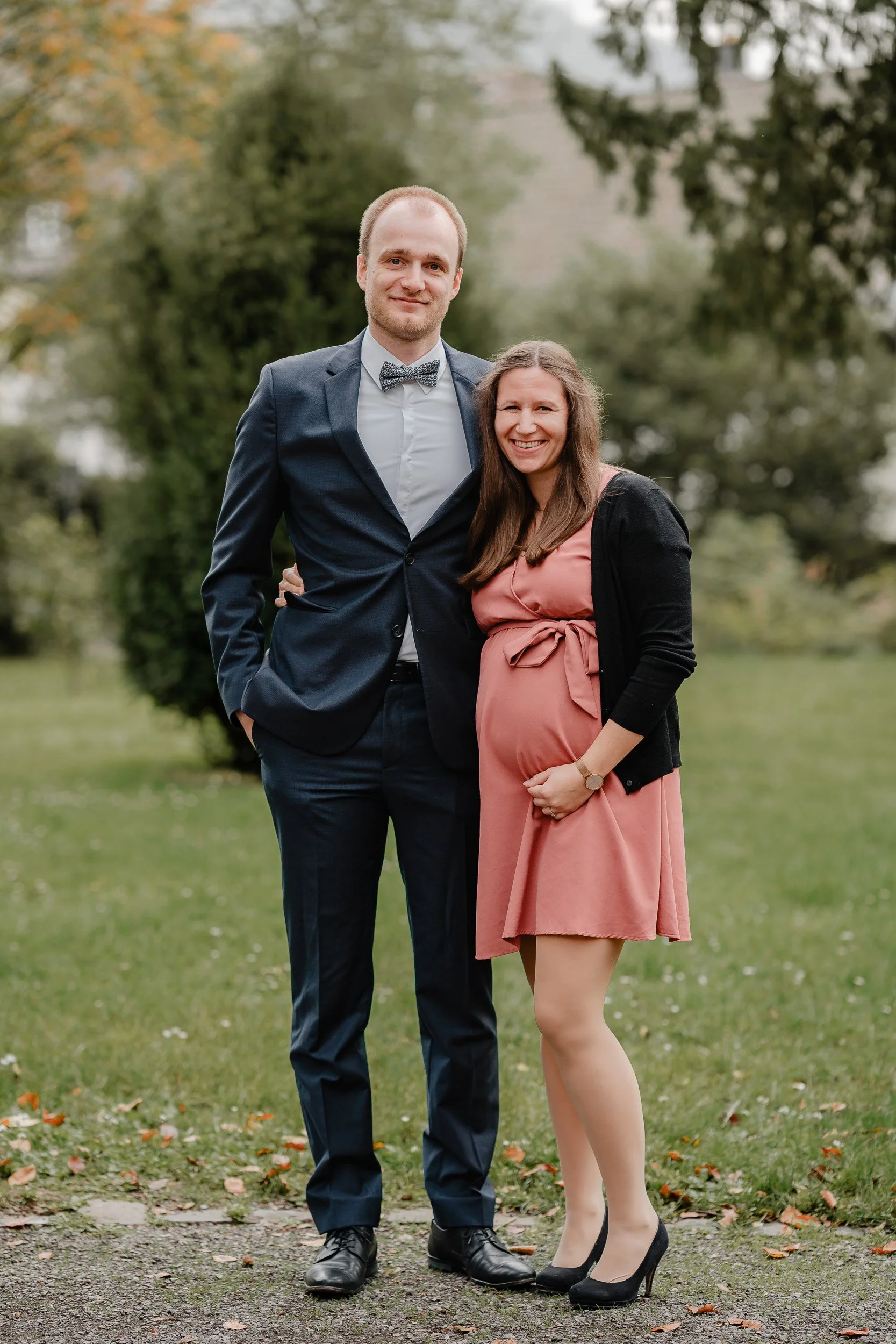 Ein Mann in Anzug mit Fliege, eine schwangere Frau in einem pinken Kleid mit schwarzer Jacke und High Heels, stehen auf einer Wiese in einem Park.