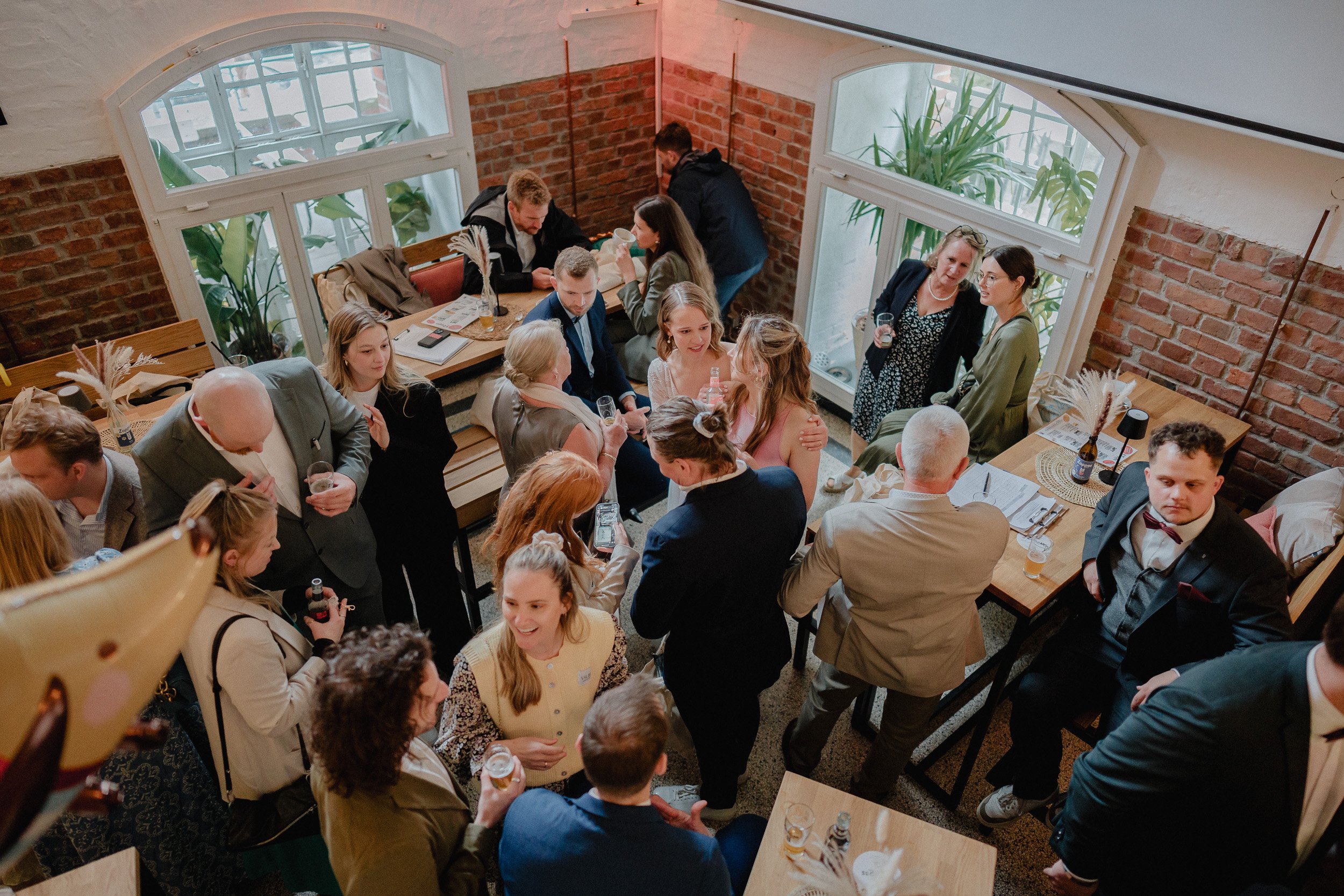 Menschen feiern in einem rustikalen Raum mit Ziegelwänden, einige sitzen, andere stehen, trinken und unterhalten sich.
