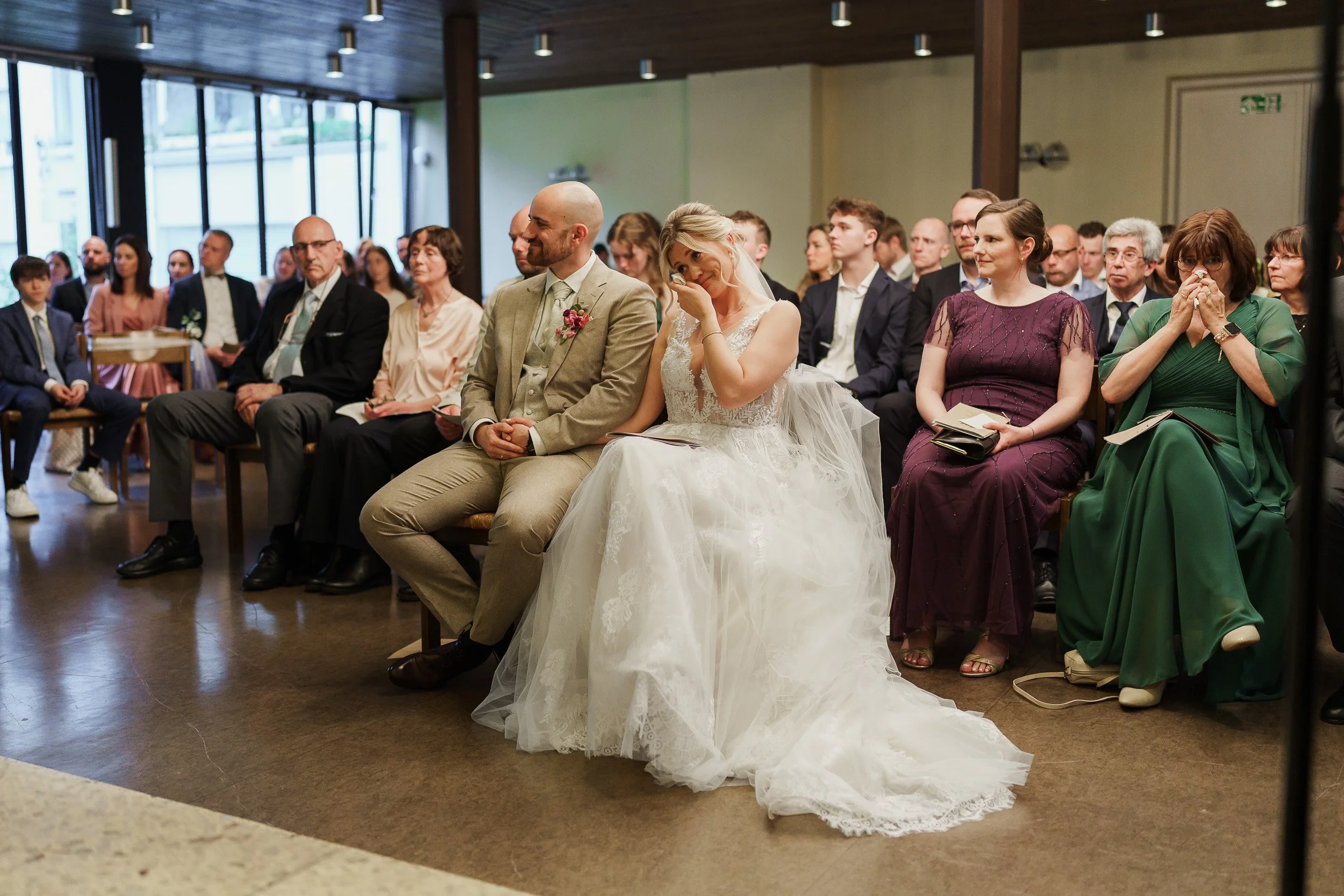Hochzeitspaar sitzt während der Zeremonie vor versammelter Gesellschaft in einem Saal.