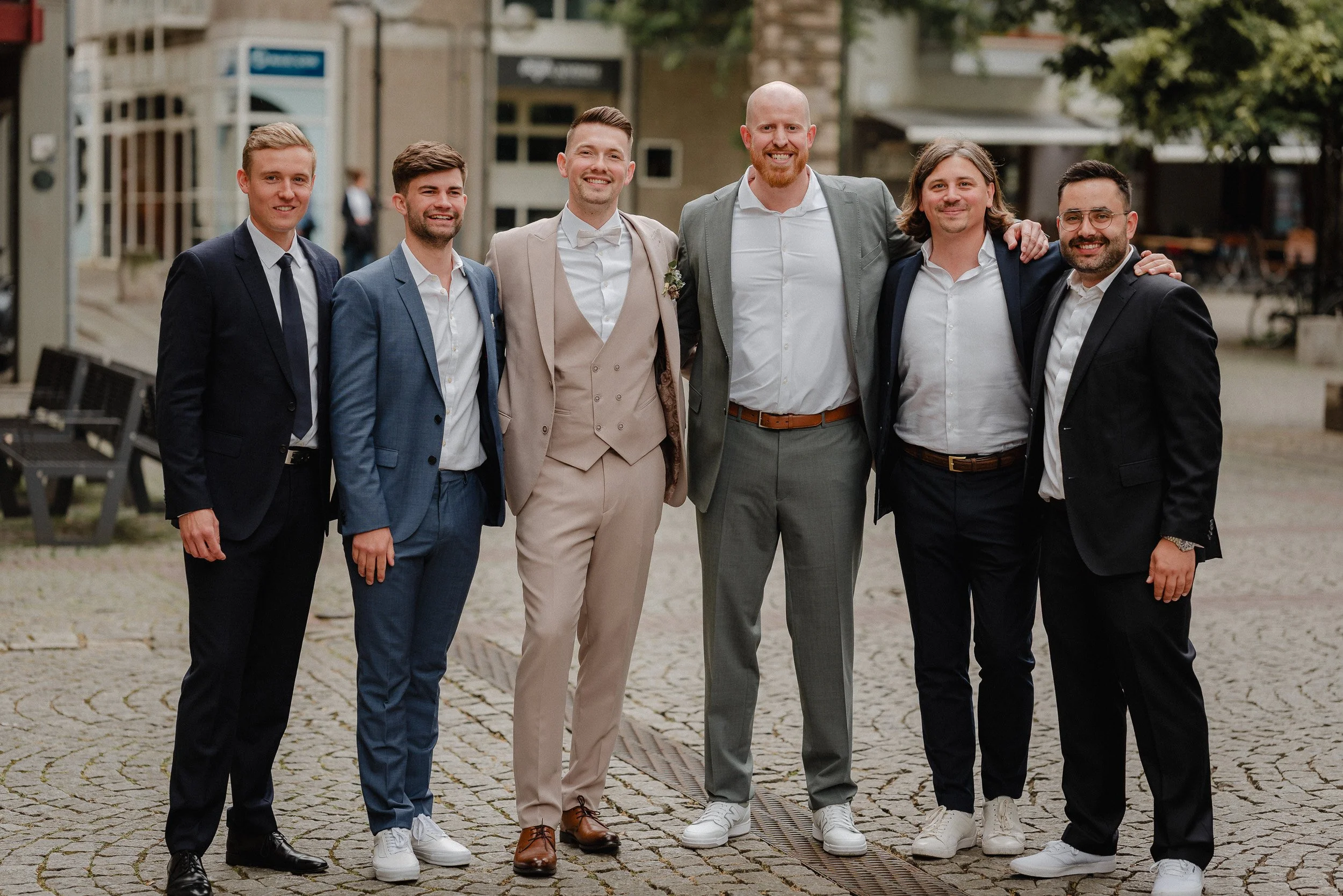 Gruppe von sieben jungen Männern in Anzügen, die arm in arm auf einer gepflasterten Plaza stehen, bei einer formellen Gelegenheit, mit einem historischen Gebäude im Hintergrund.