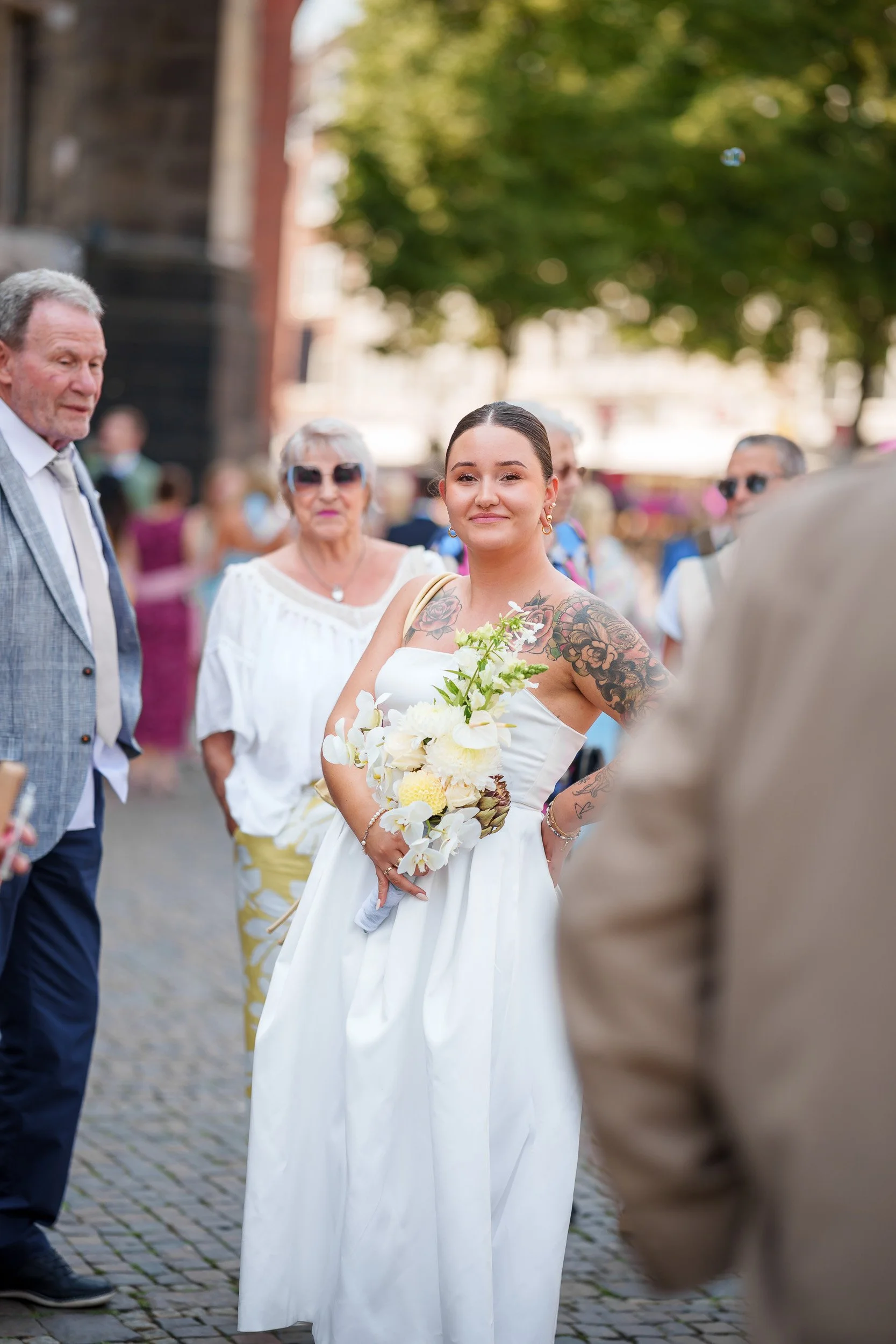 Eine junge Frau trägt ein weißes Hochzeitskleid, hält einen Blumenstrauß und steht in einer Gruppe von Menschen im Freien.