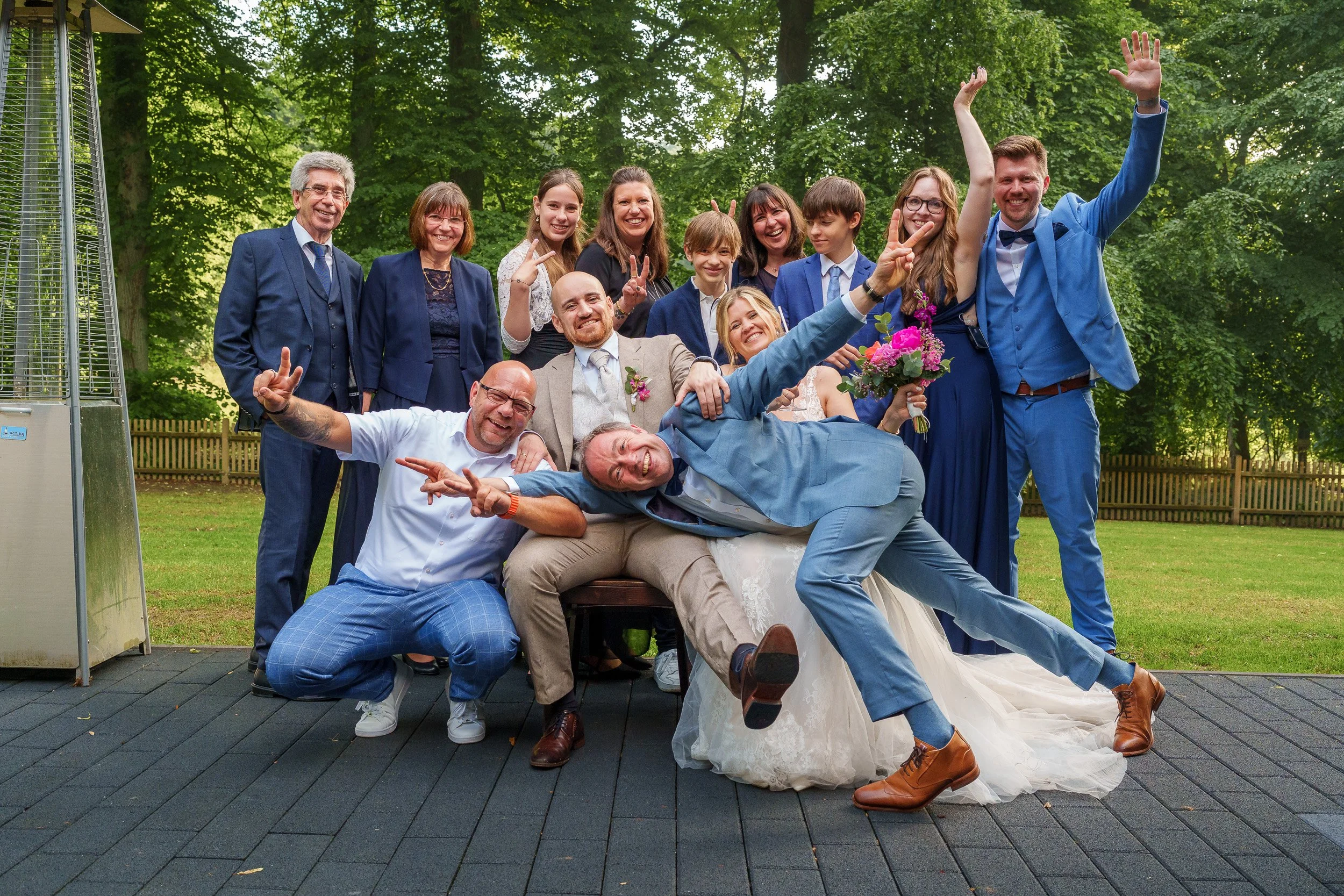 Eine fröhliche Gruppe von Menschen bei einer Hochzeit im Freien, einige lachen und posieren ausgelassen, in formeller Kleidung, umgeben von grünen Bäumen.