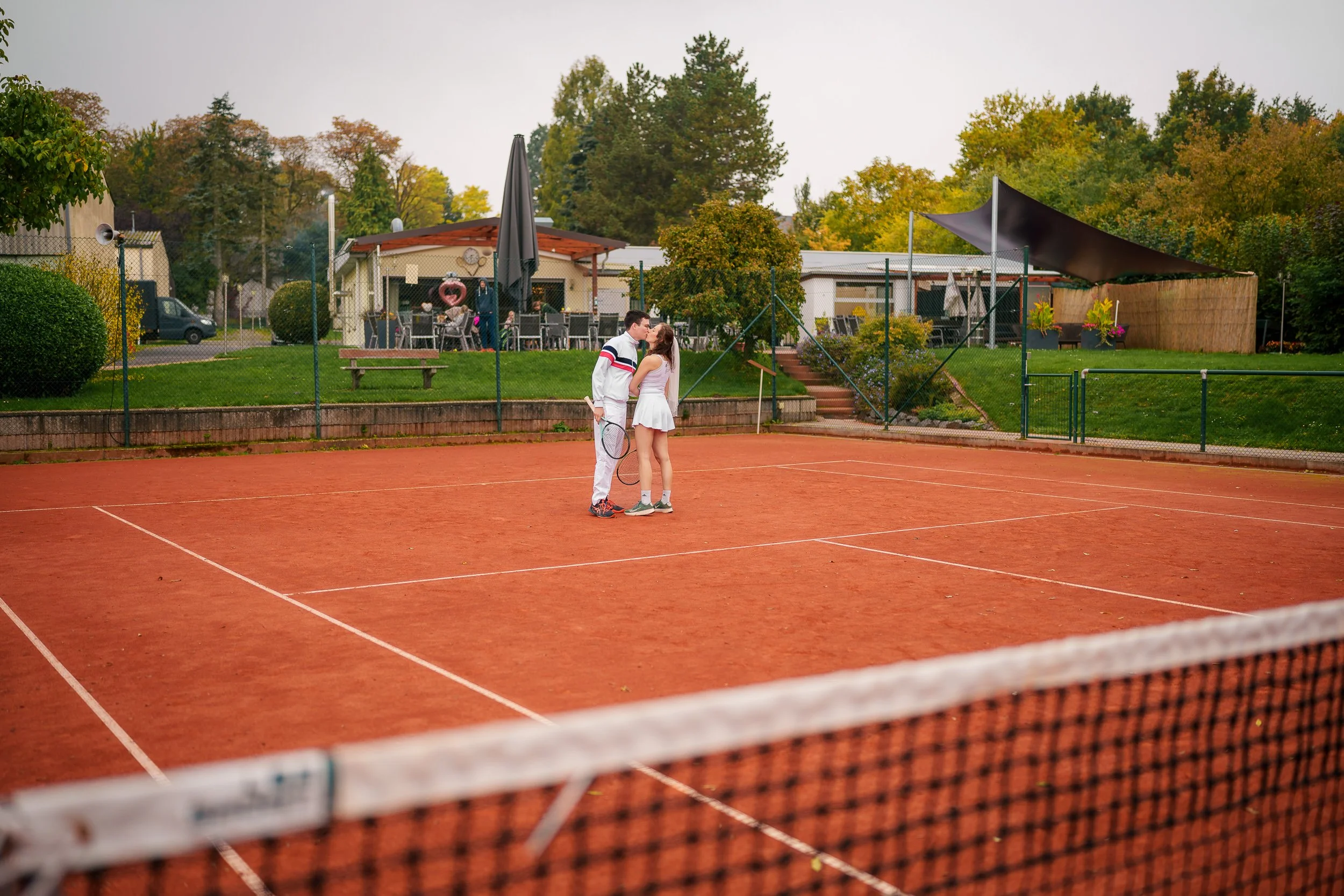 Ein Tennisplatz im Freien mit einem jungen Paar, das sich beim Tennis küsst, im Hintergrund Gäste beim Essen im Garten eines Hauses, umgeben von Bäumen und bewölktem Himmel