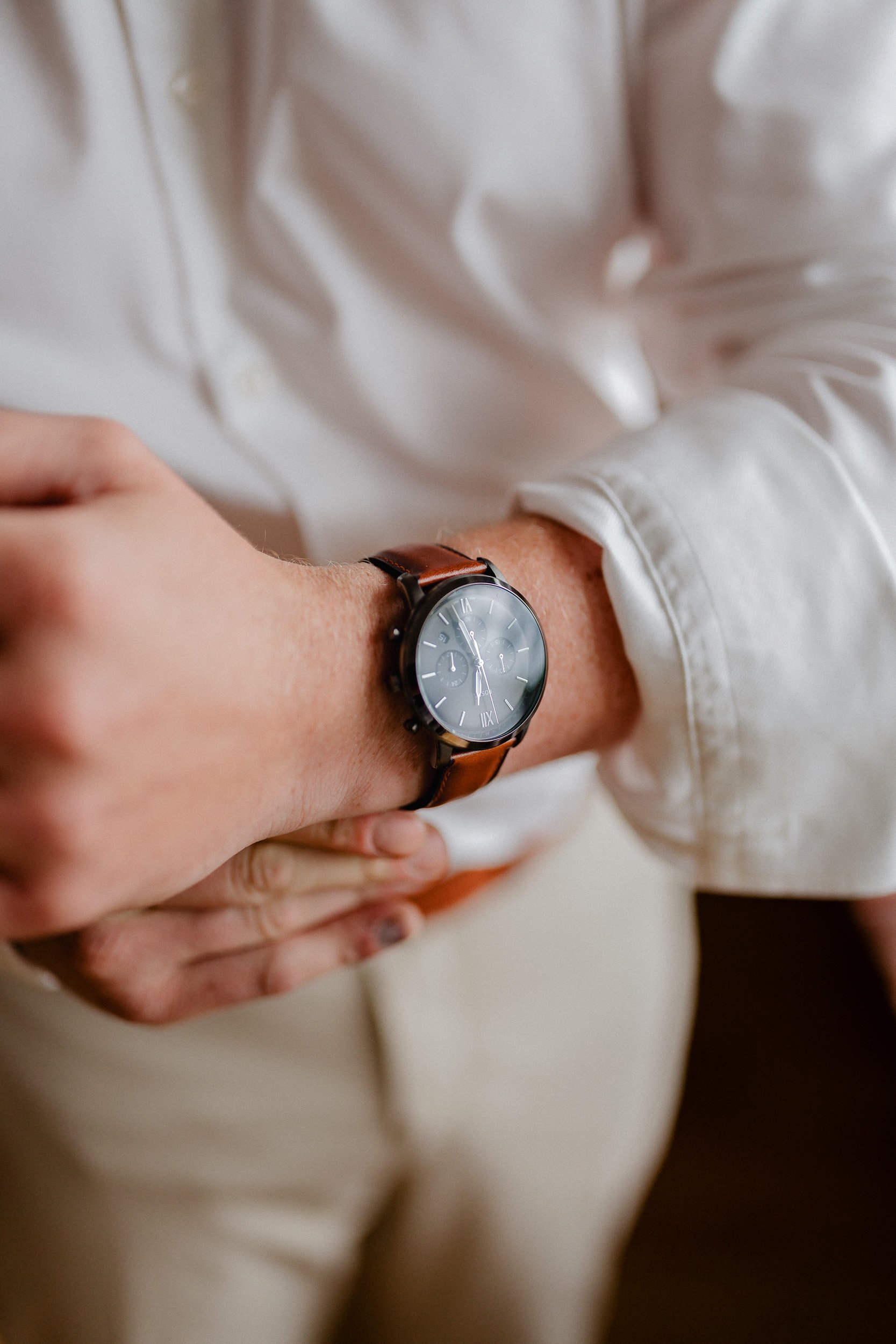 Nahaufnahme einer Person mit einem Armbanduhr mit braunem Lederarmband, die Hand liegt auf dem Bauch, Person trägt ein weißes Hemd.