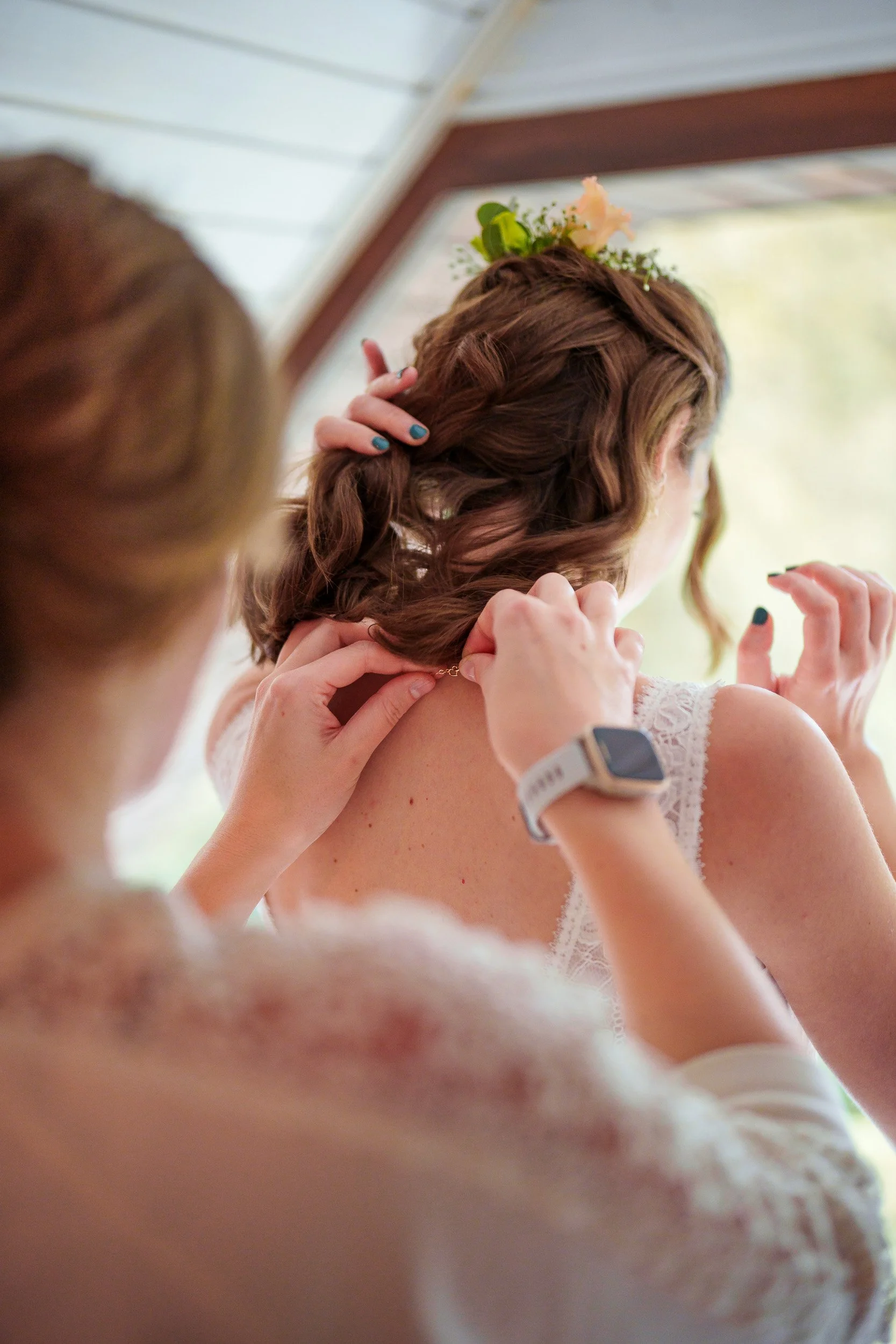 Eine Frau wird von einer anderen Person dabei geholfen, sich ein Schmuckstück an den Hals zu legen, vermutlich bei einer Hochzeitszeremonie. Die Frau trägt ein weißes Kleid und hat braune, wellige Haare mit Blumen geschmückt.
