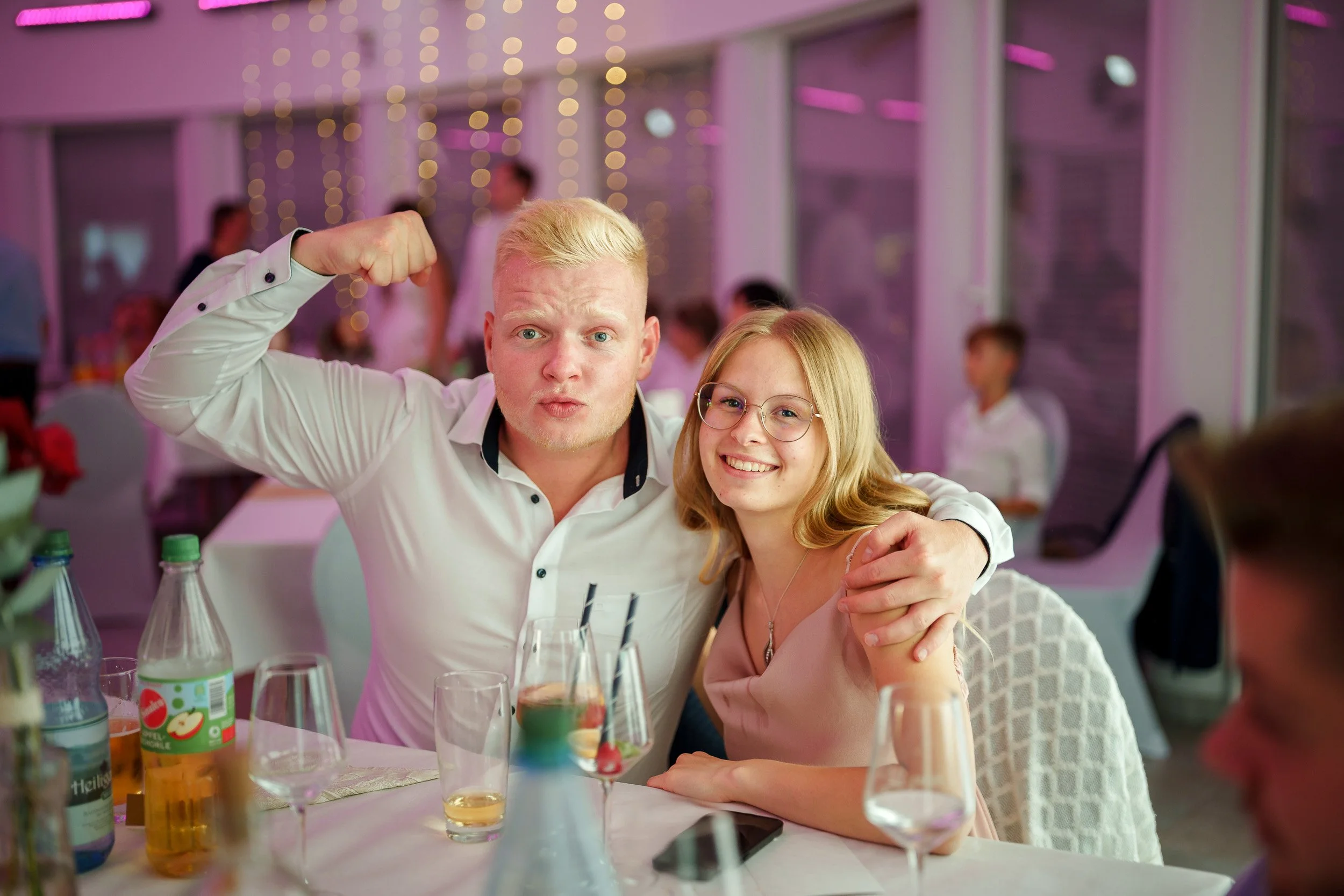 Ein lachendes junges Mädchen mit Brille sitzt neben einem jungen Mann, beide lächelnd bei einer Feier zwischen Getränken auf einem festlich gedeckten Tisch.