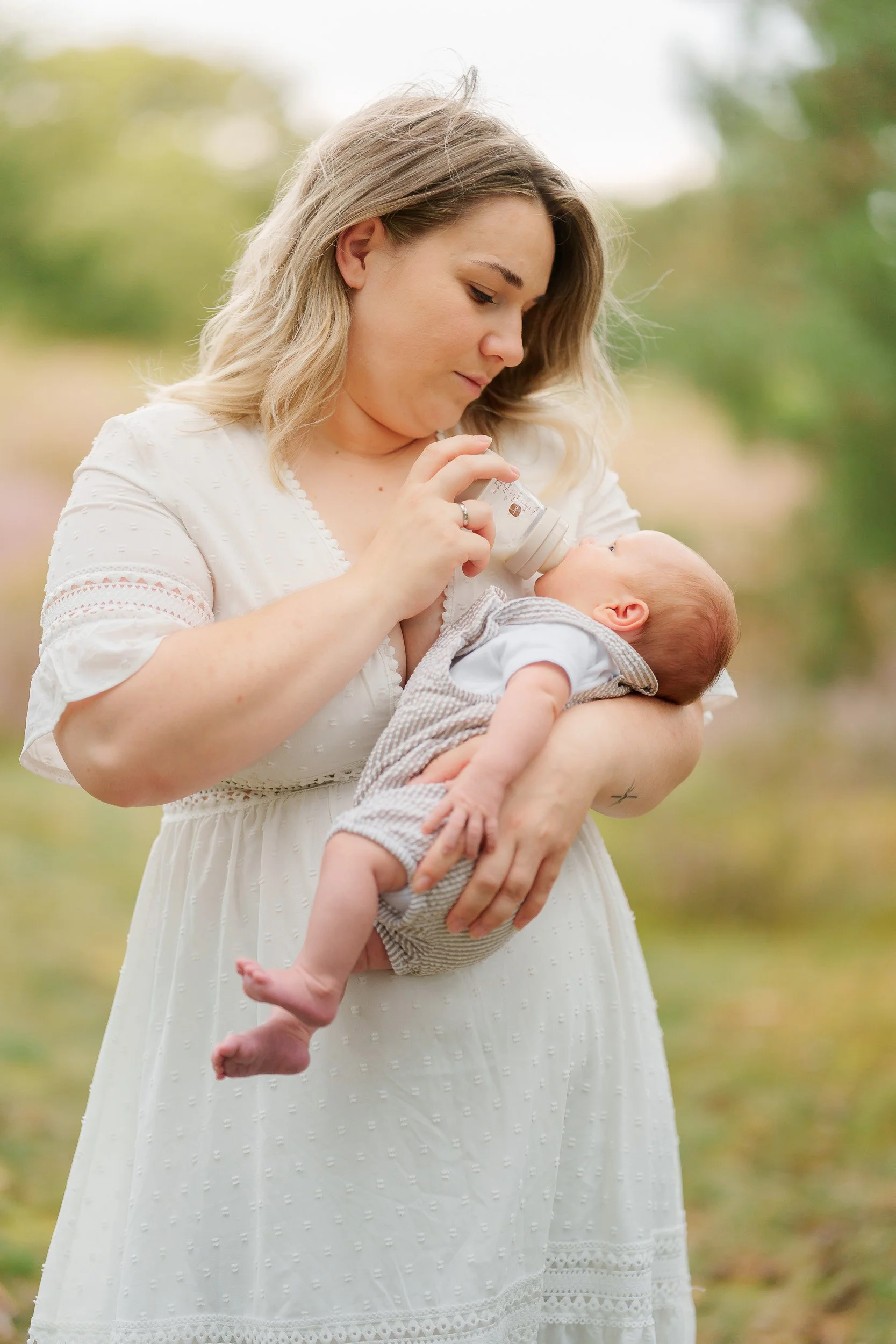 Eine Frau in einem weißen Kleid hält ein Baby im Arm und gibt ihm bei einem Fläschchen Milch.