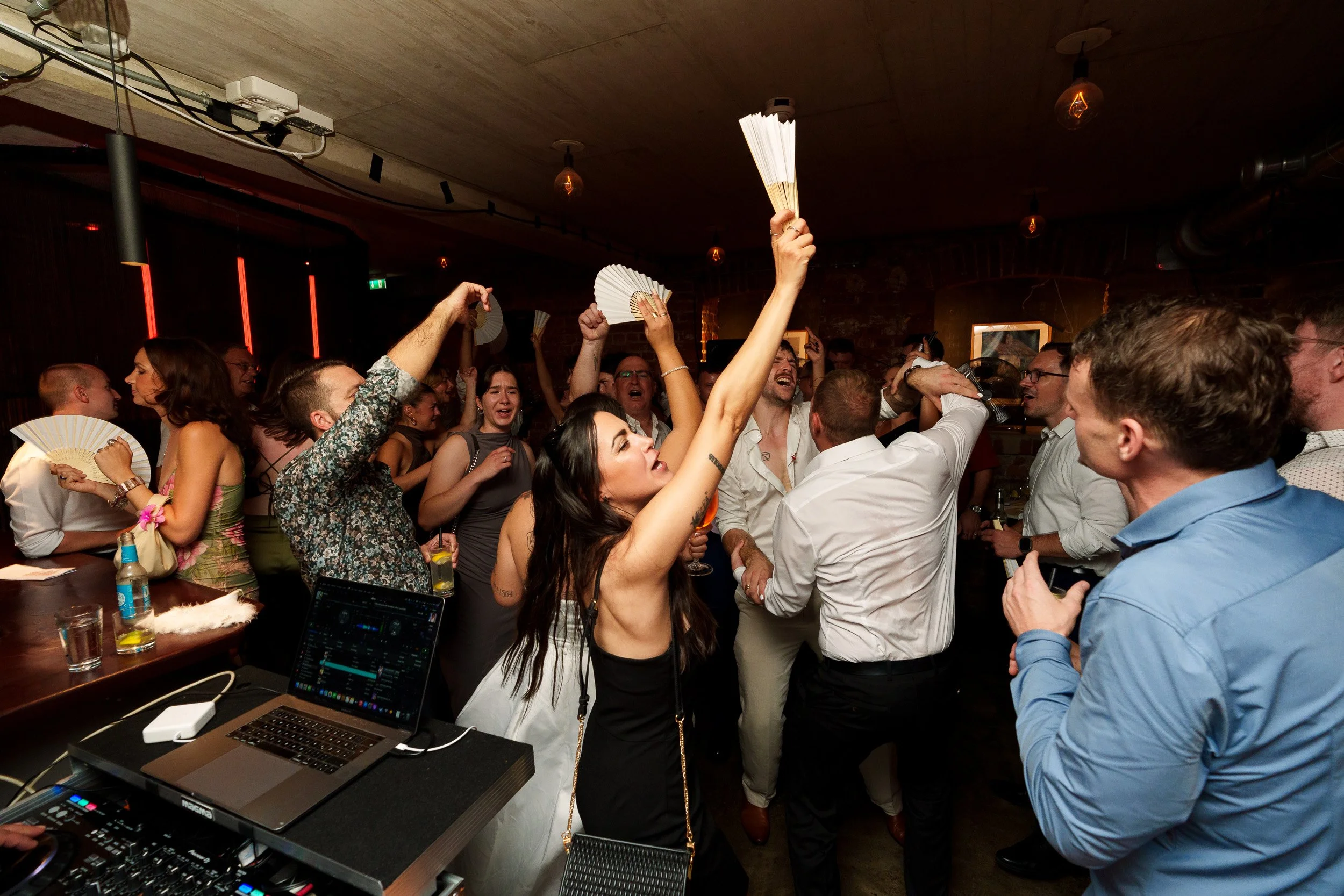 Menschen feiern und tanzen in einer Bar, einige halten Fächer hoch, die Musik wird von einem DJ gespielt, auf einem Tisch stehen Getränke.