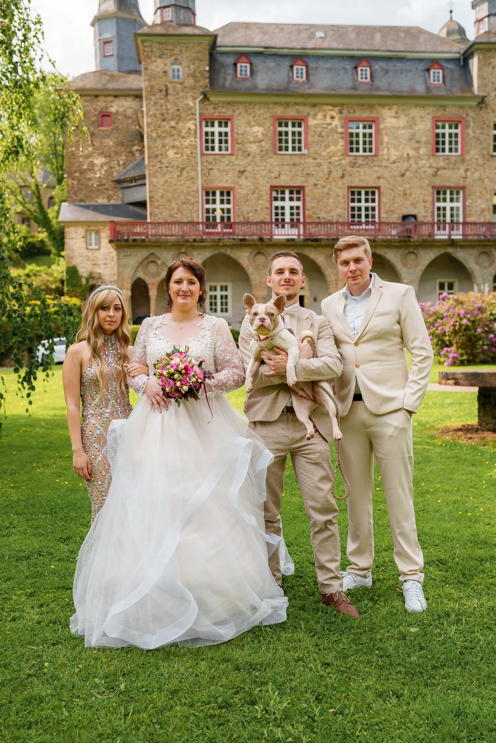 Gruppe von fünf Menschen und einem Hund vor einem historischen Gebäude im Garten, bei einer Hochzeit.