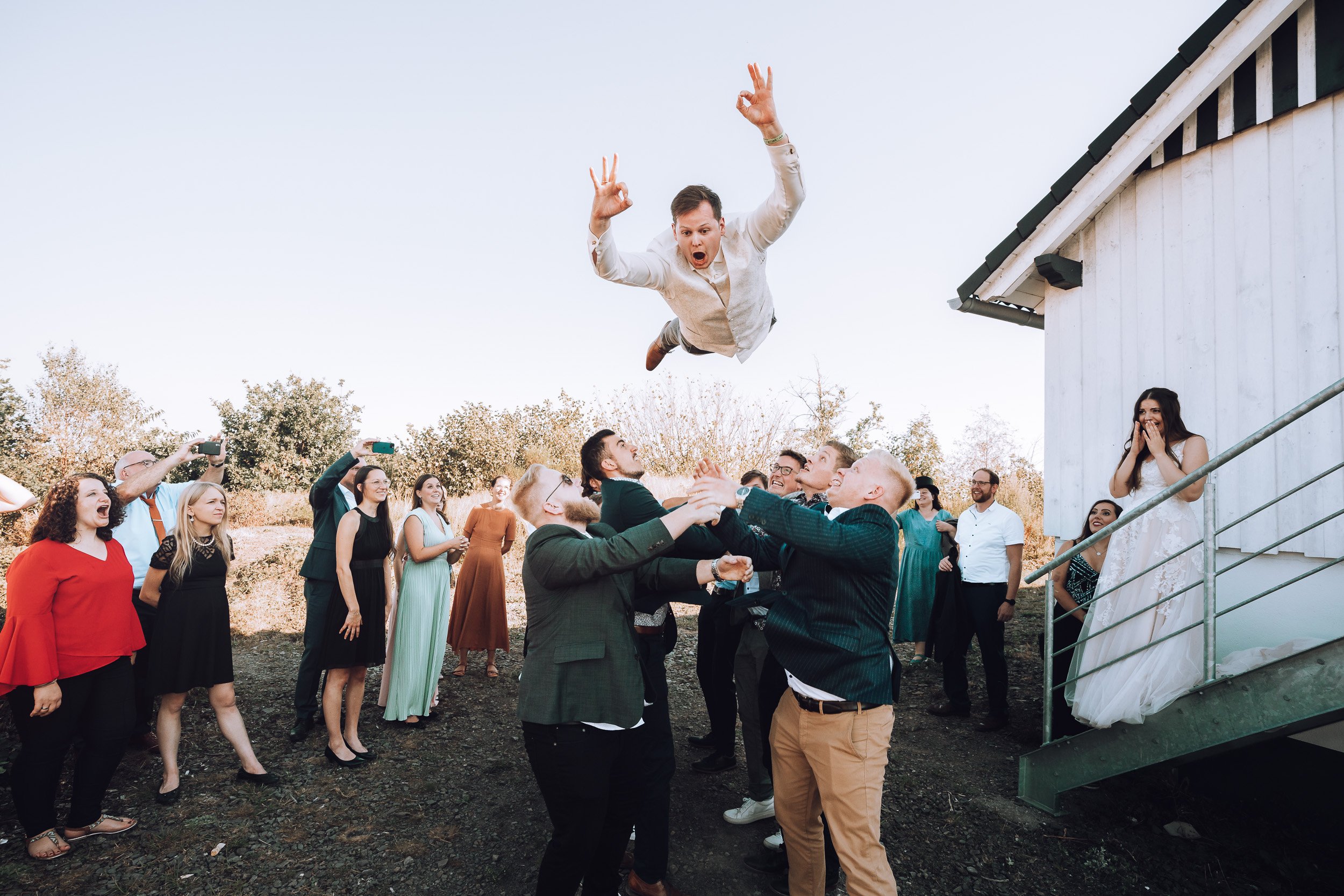 Eine Gruppe von Menschen beim Hochzeitsfest, die eine Person in die Luft heben, während eine Braut auf der Eingangstreppe steht und alle lachen und Fotos machen.