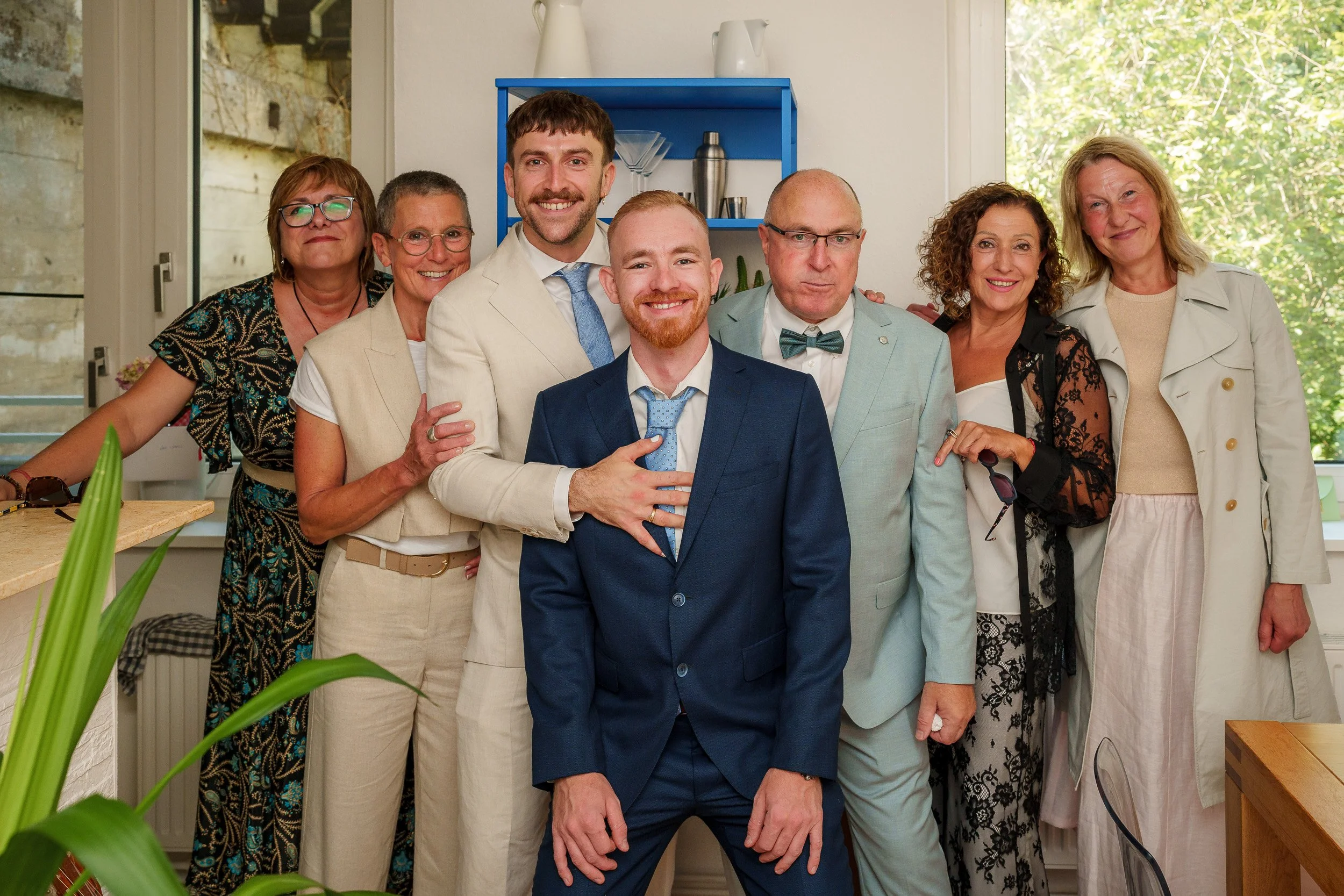 Gruppe von acht Menschen, die bei einer Feier in einem Wohnzimmer lachen und posieren, einige tragen formelle Kleidung.