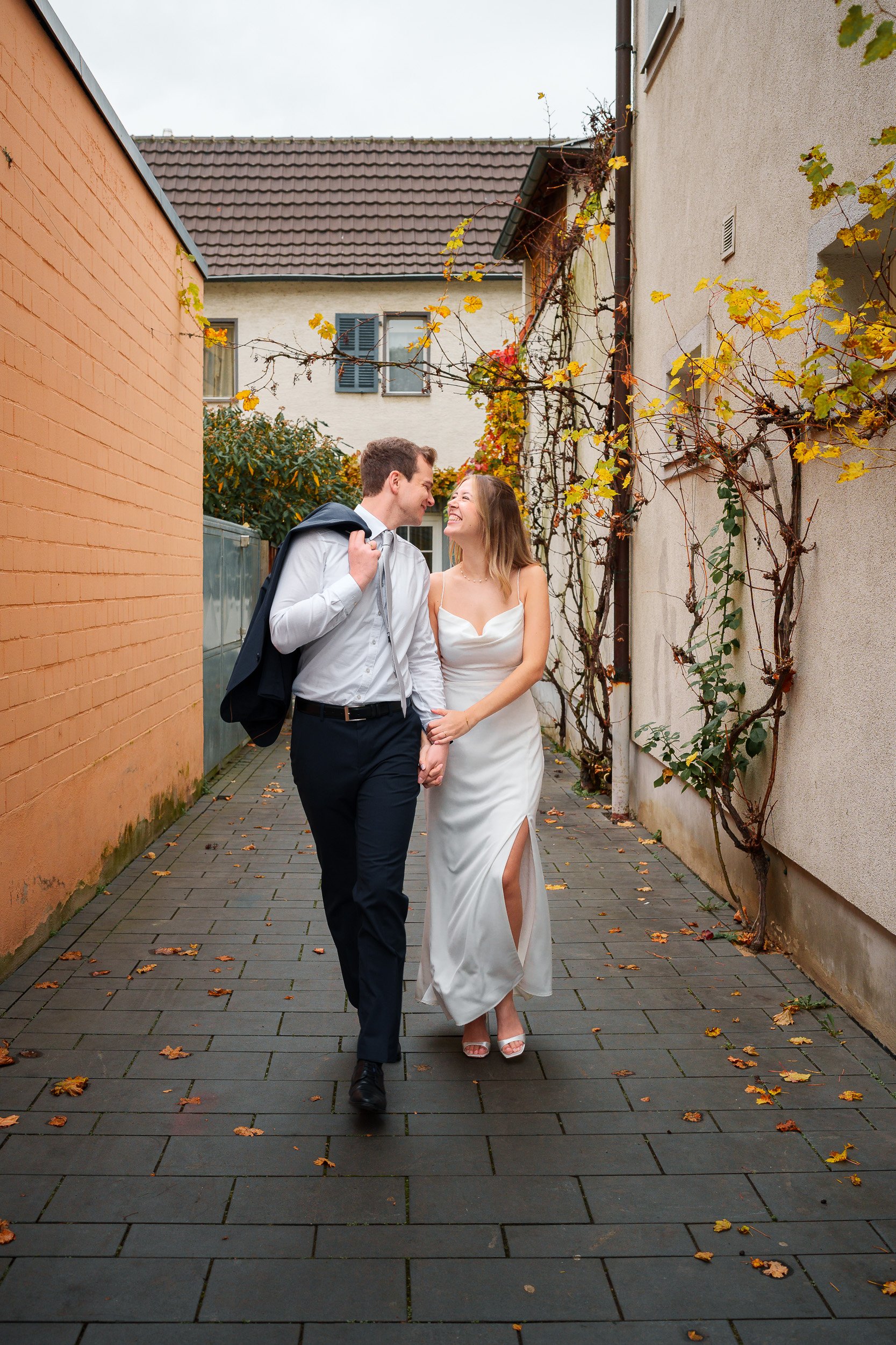 Ein junges Paar, das einander Händchen haltend durch einen engen, mit Laub bedeckten Weg geht. Sie tragen elegante Kleidung, sie in einem weißen Kleid, er in einem weißen Hemd und dunklen Hosen, mit einem Jackett über seiner Schulter. Sie lachen und 