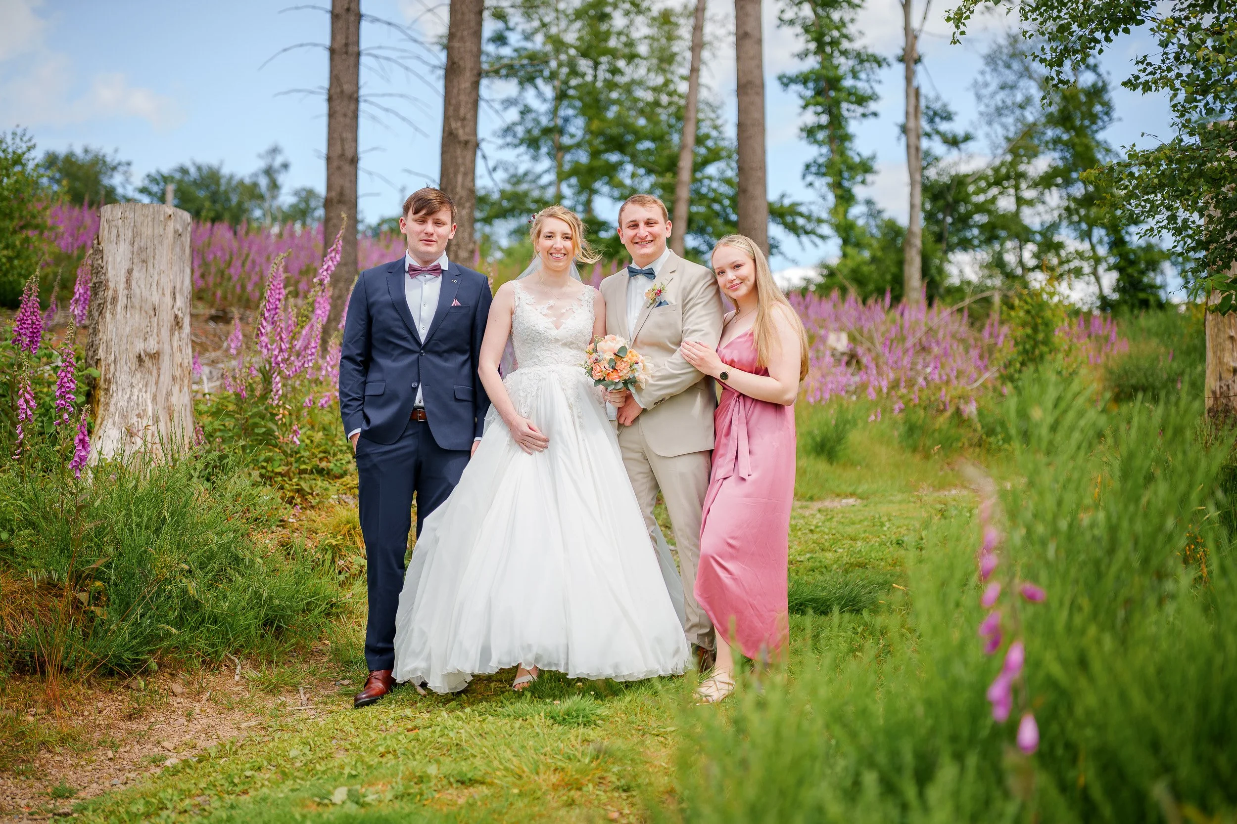 Eine Hochzeitsgruppe aus fünf Personen, bei einer Hochzeit im Freien mit Bäumen und lila Blumen im Hintergrund.