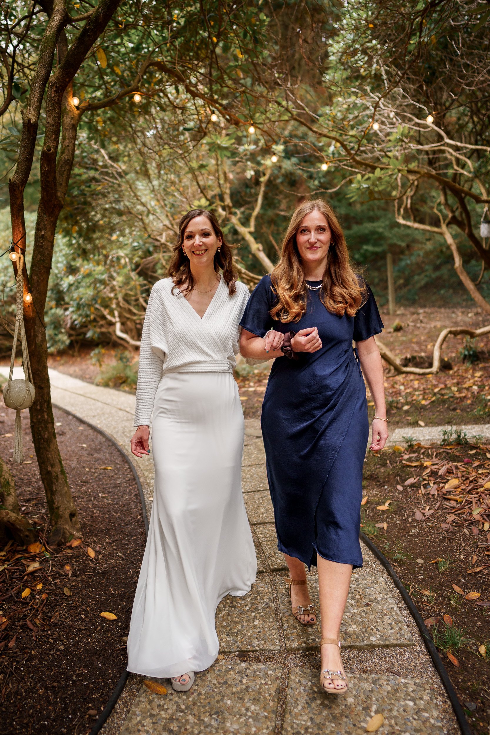 Zwei Frauen, eine in einem weißen Brautkleid und die andere in einem dunkelblauen Kleid, gehen Arm in Arm auf einem Gartenweg, umgeben von Bäumen und Lichterketten, bei einer Feier oder Hochzeit.
