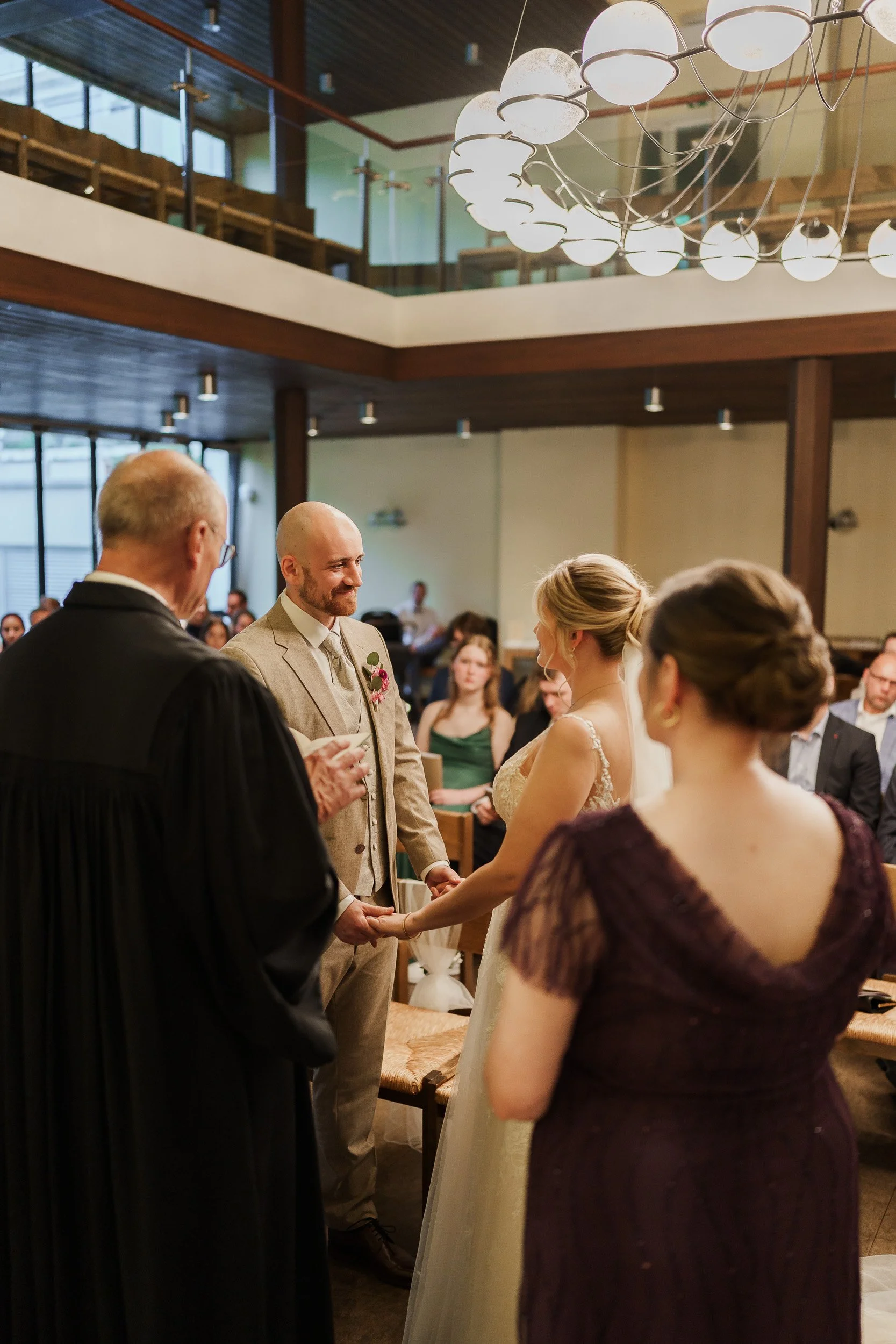 Trauung bei einer Hochzeit in einer modernen Kirche, Braut und Bräutigam halten Händchen, Pfarrer spricht, Gäste im Hintergrund.