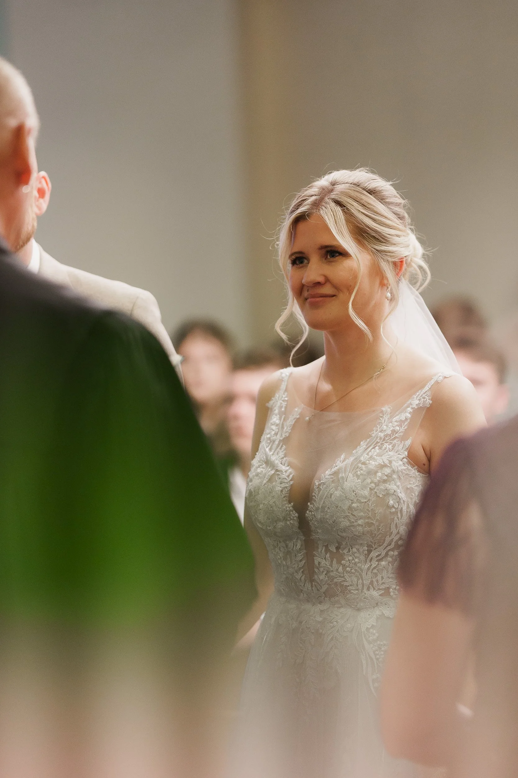 Braut mit lockigem, schulterlangem Haar und weißem, spitzenbesticktem Brautkleid bei einer Hochzeitszeremonie.