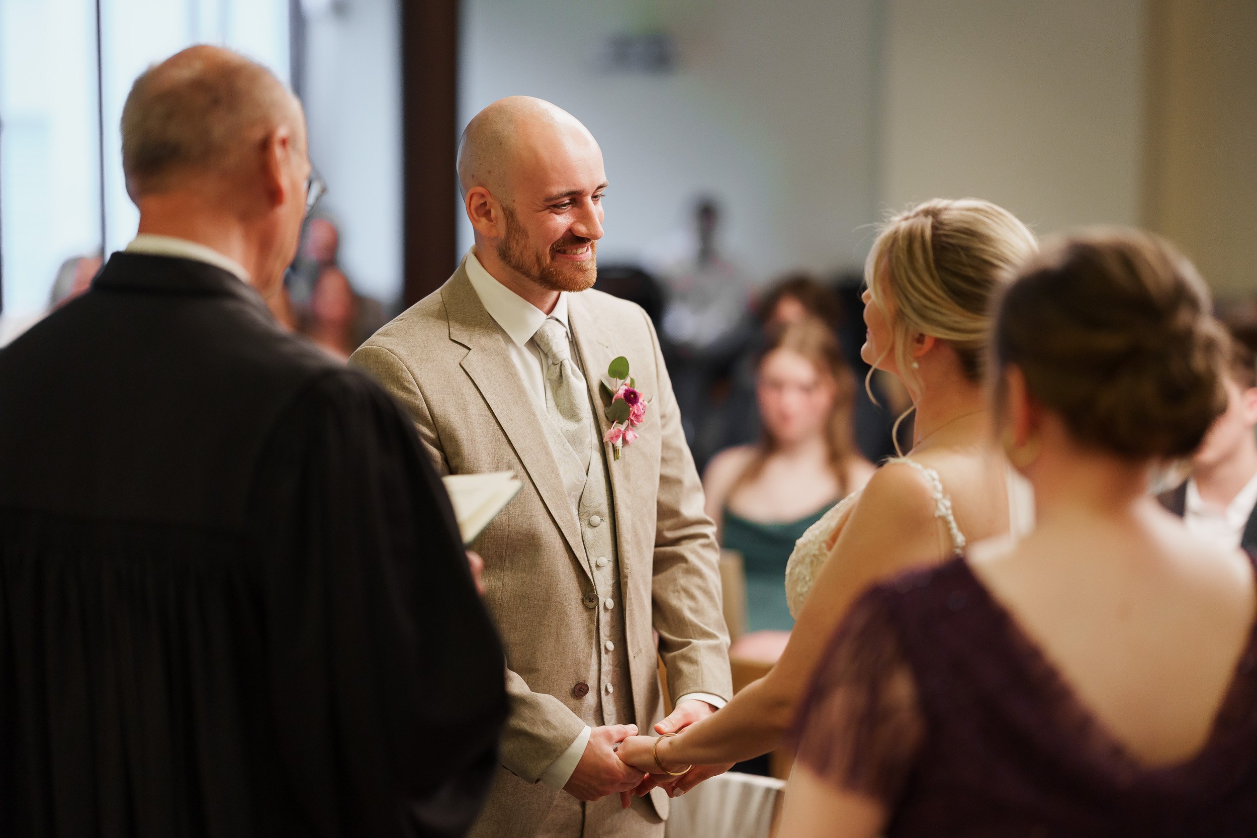 Ein Mann und eine Frau bei ihrer Hochzeitszeremonie, die sich die Hände halten, während ein Trauredner das Paar segnet, im Hintergrund zahlreiche Gäste.