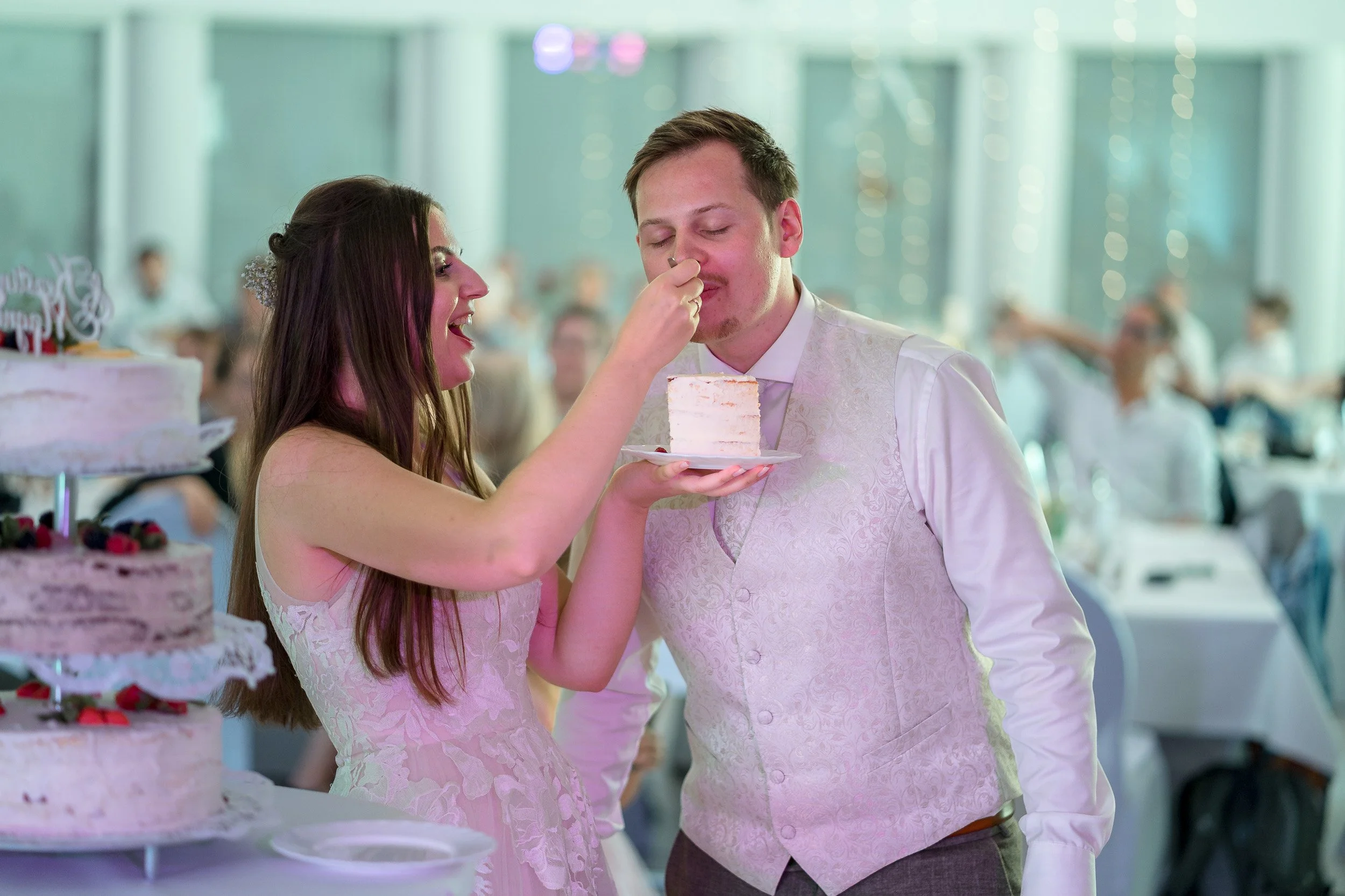 Ein Brautpaar bei einer Hochzeit, sie füttert den Bräutigam mit einem Stück Hochzeitstorte. Im Hintergrund sind weitere Hochzeitsgäste und ein mehrstöckiger Hochzeitstorte. Die Atmosphäre ist festlich und fröhlich.