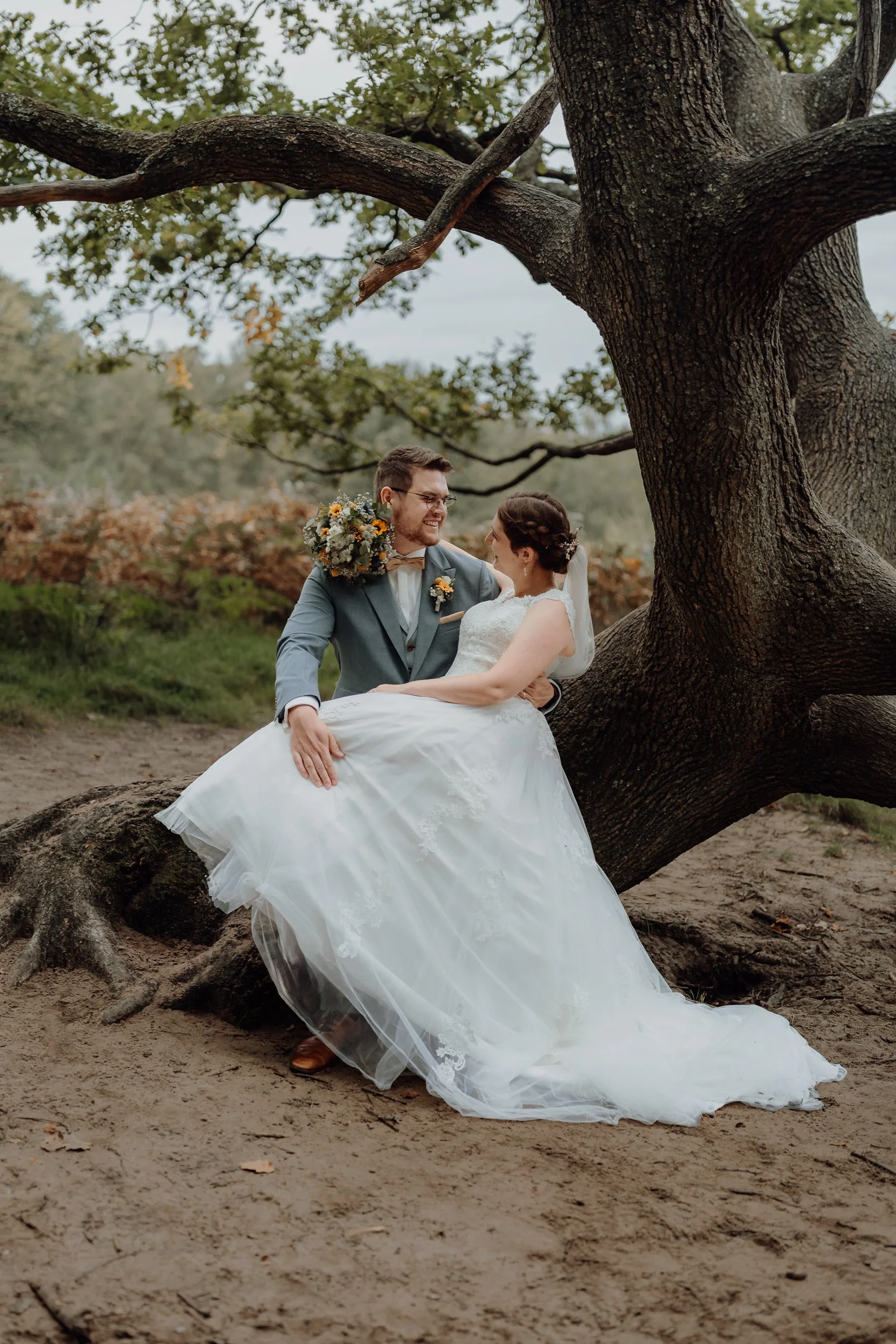 Ein Brautpaar sitzt entspannt an einem Baum im Grünen. Der Bräutigam trägt einen blauen Anzug mit weißem Hemd und Krawatte, die Braut trägt ein weißes Hochzeitskleid mit Spitzendetails. Der Bräutigam hält die Braut lachend in den Armen. Die Szene ist
