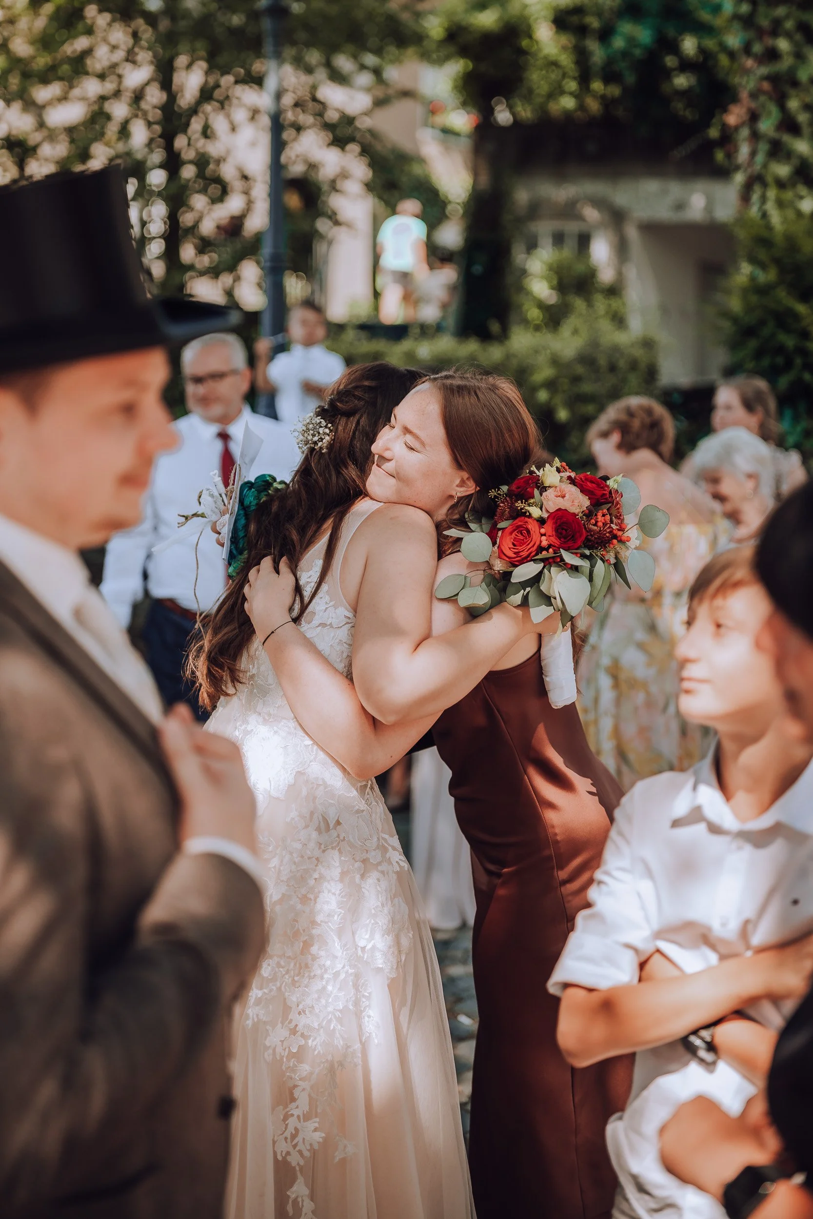 Zwei Frauen bei einer Hochzeit umarmen sich, eine trägt ein weißes Brautkleid mit Spitzen und hält einen Blumenstrauß, die andere trägt ein braunes Kleid.