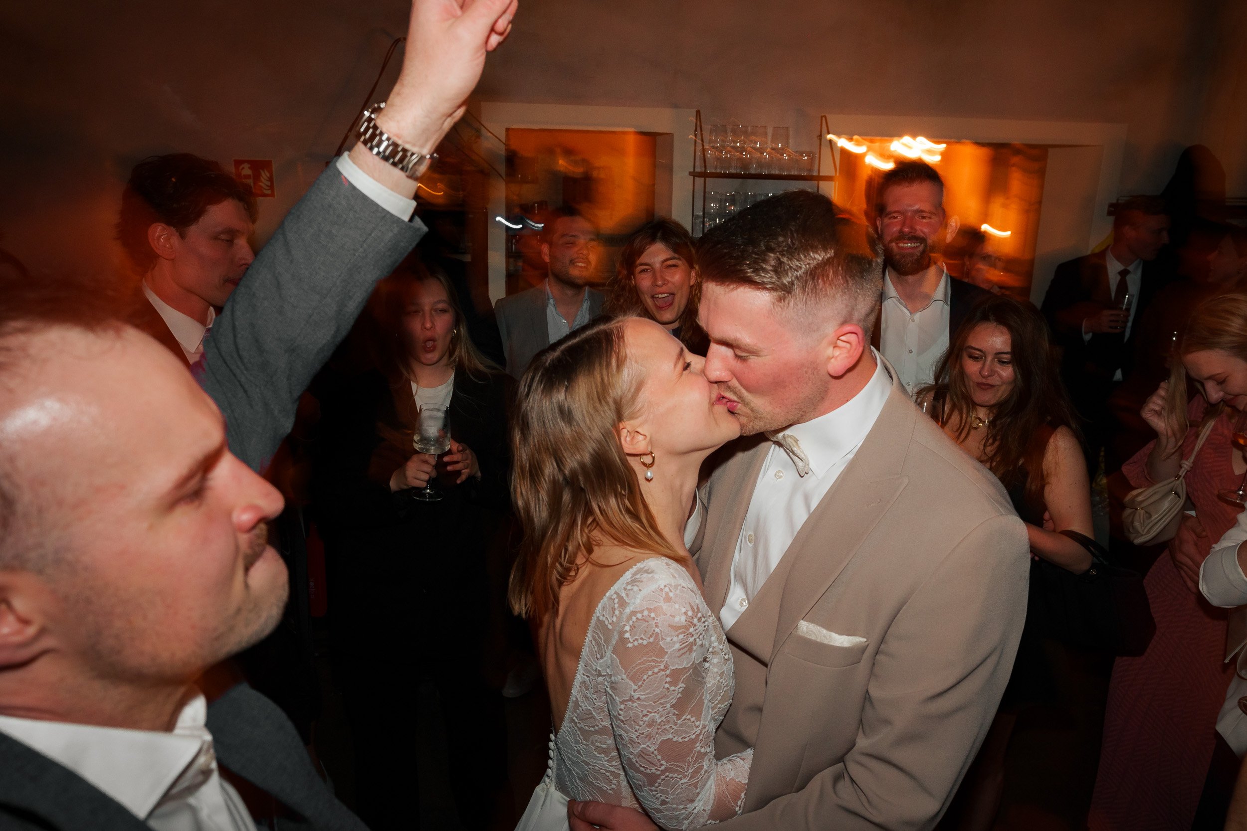 Ein Brautpaar küsst sich inmitten einer Hochzeitsgesellschaft bei einer Feier, die Menschen um sie herum sind fröhlich und feiern.