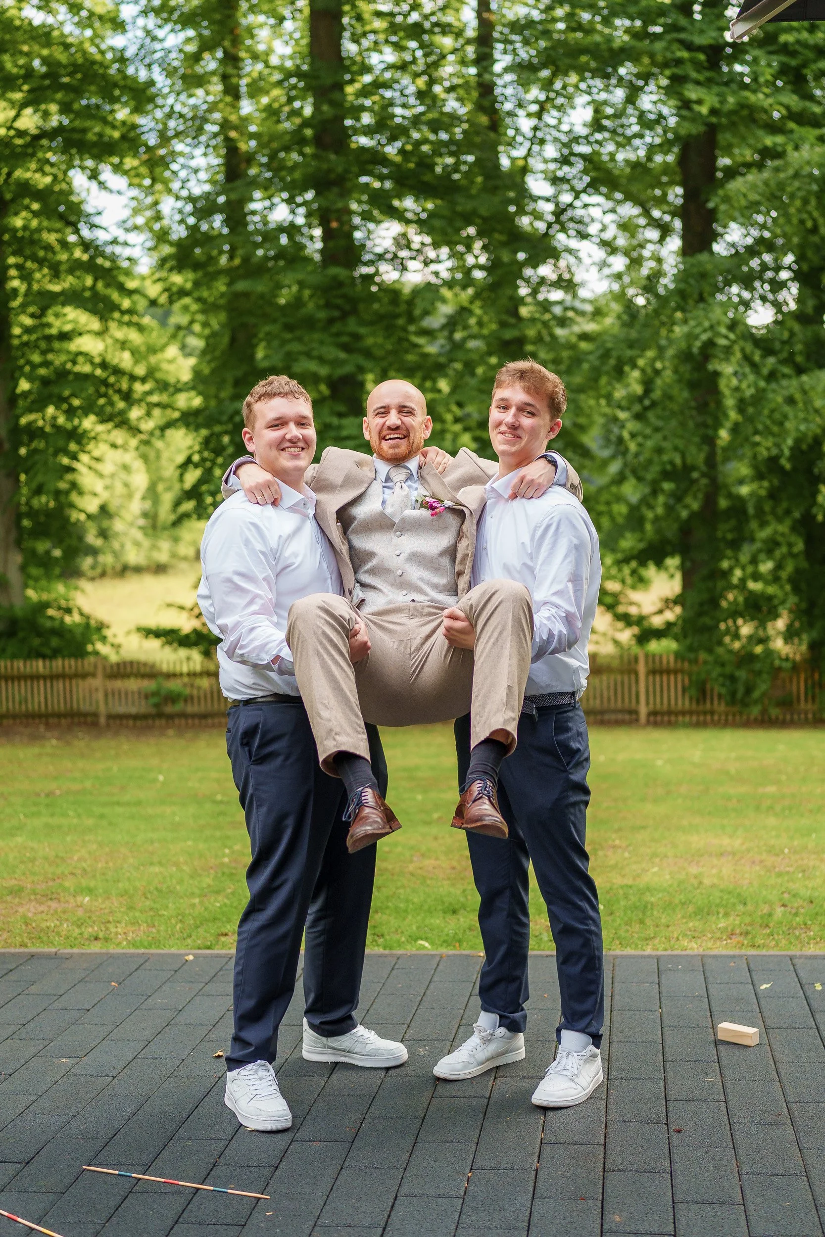Drei junge Männer in formeller Kleidung, einer in der Mitte trägt einen Anzug und ein Blumenanstecker, wird von den beiden anderen Männern getragen, während sie lachen und Spaß im Freien haben.