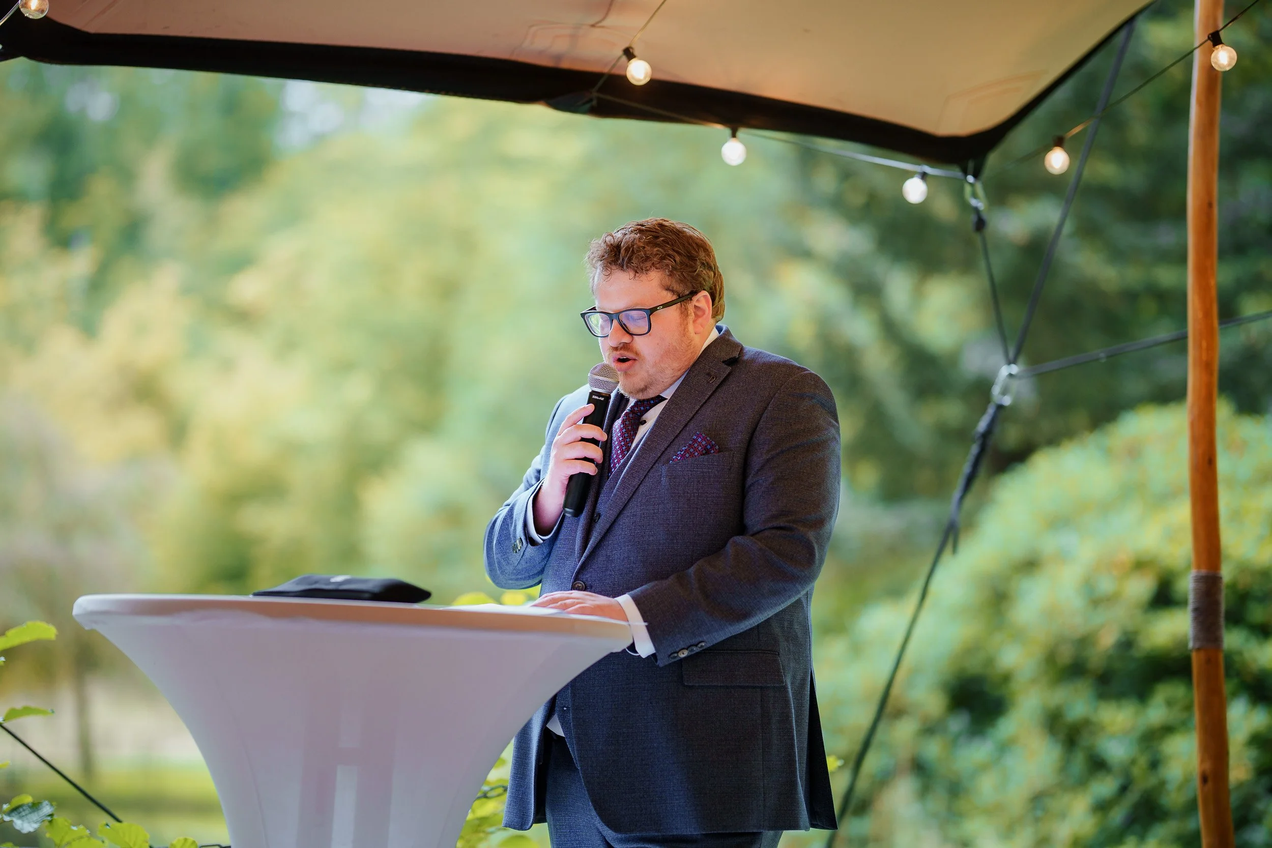 Ein Mann in einem grauen Anzug hält eine Handmikrofon und spricht bei einer Outdoor-Veranstaltung unter einem Zelt mit Lichterketten, im Hintergrund sind Bäume und grünes Laub.