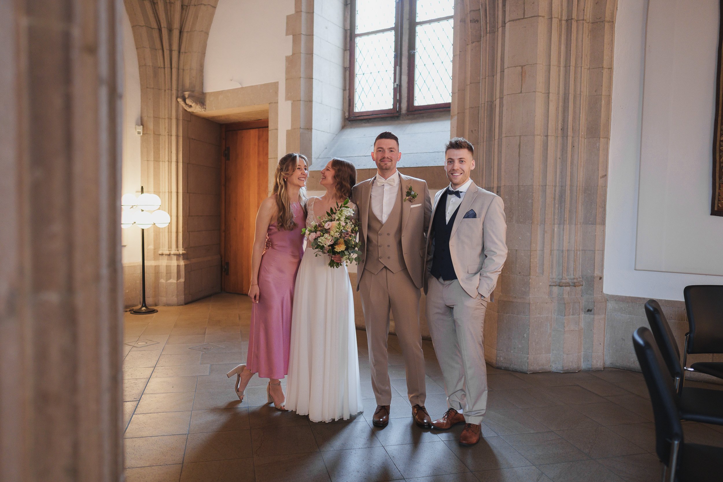 Eine Hochzeitsgruppe bestehend aus vier Personen, zwei Frauen und zwei Männer, steht in einem hellen Raum mit architektonischen Elementen. Die Braut trägt ein weißes Kleid und hält einen Blumenstrauß, während die anderen in eleganten Anzügen oder Kle