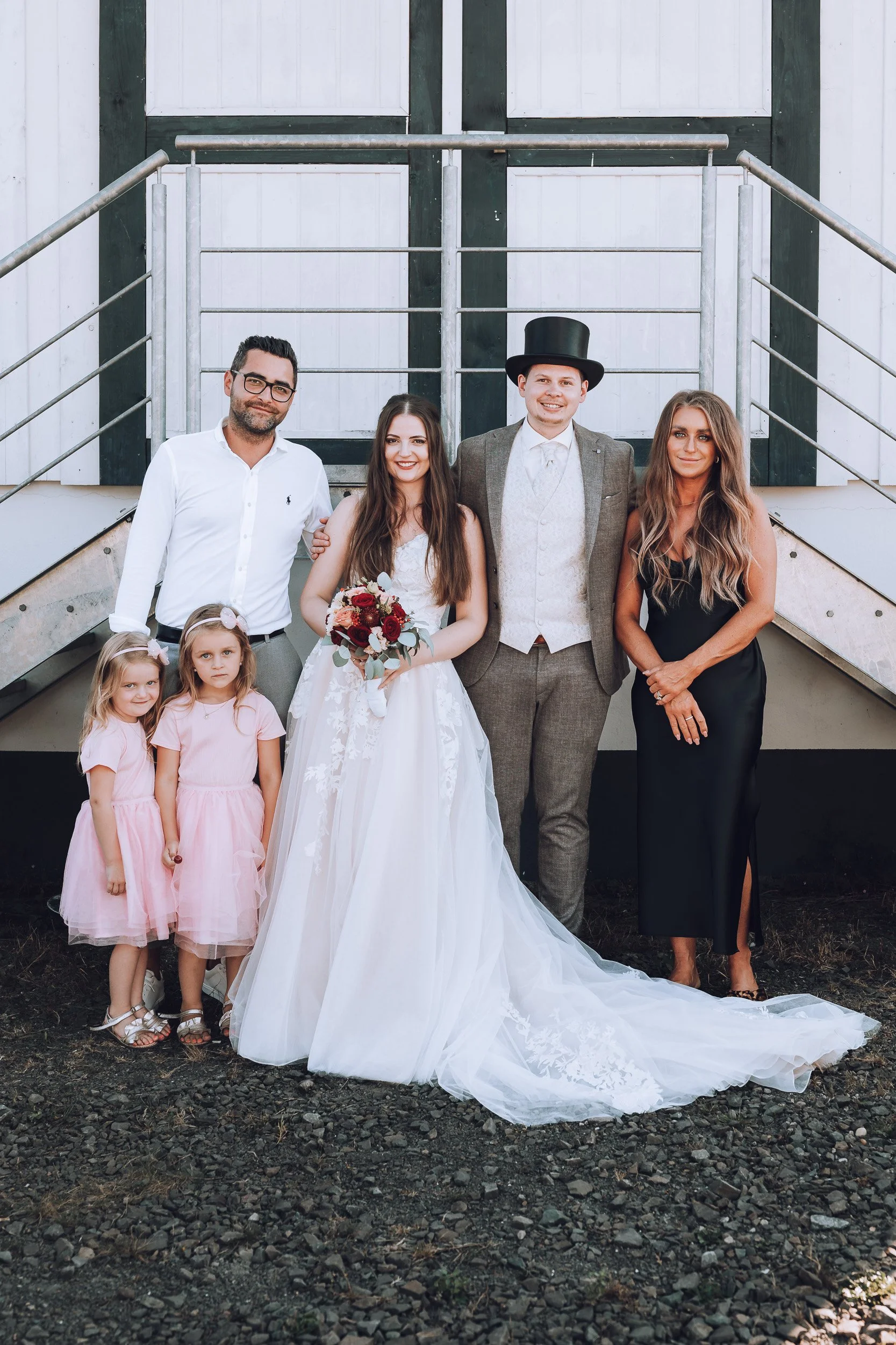Gruppe von sechs Personen, darunter Braut in Hochzeitskleid, Bräutigam mit Zylinder, zwei Frauen und zwei kleine Mädchen, vor einer weißen Scheunentür.