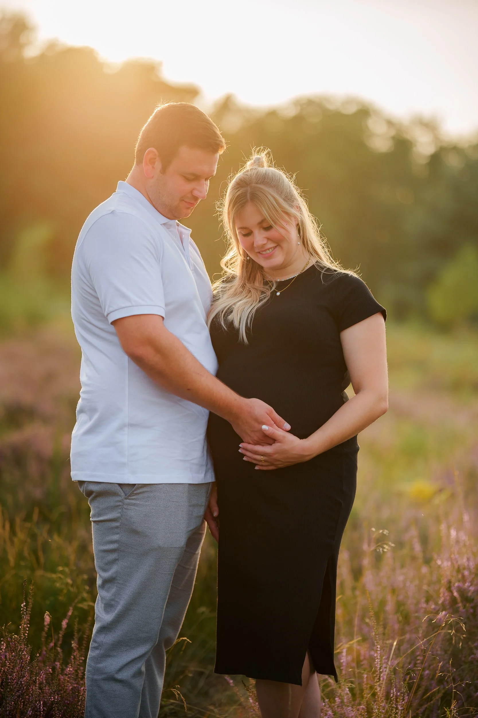 Ein Paar steht im Sonnenuntergang in einem Feld, der Mann berührt liebevoll den Bauch der schwangeren Frau.