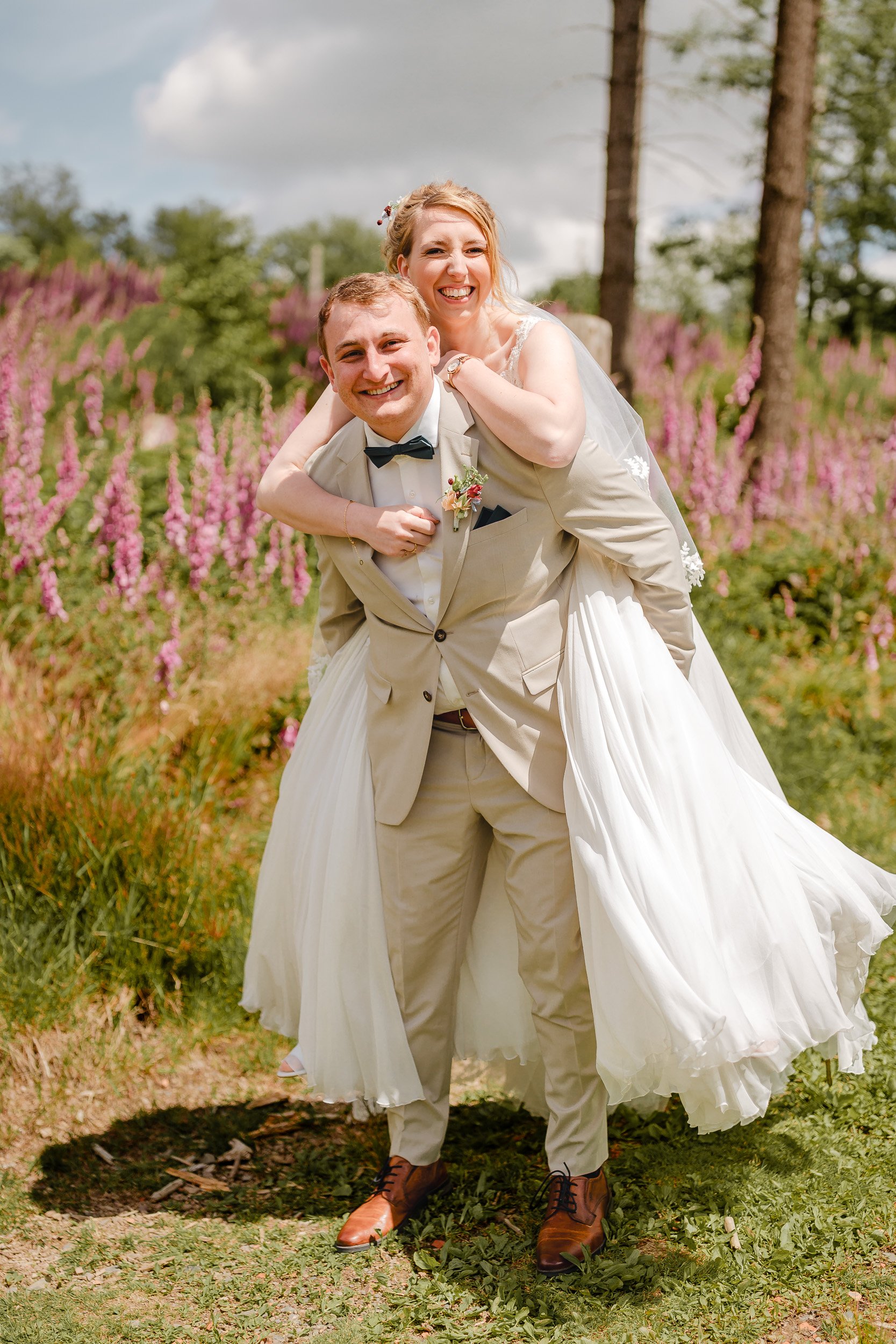 Ein lachendes Brautpaar, der Bräutigam trägt einen Mantel und Fliege, die Braut trägt ein weißes Hochzeitskleid, im Hintergrund blühende pinke Blumen und grüne Bäume.