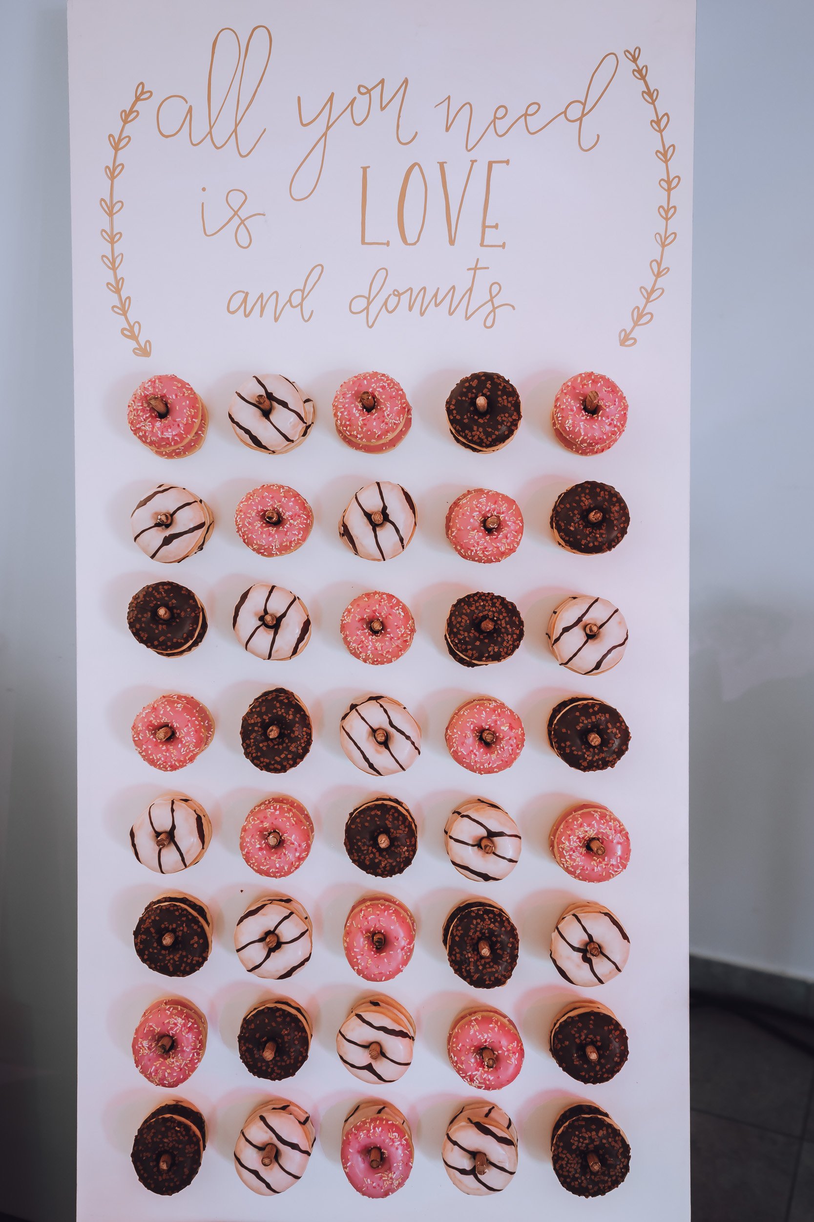 Bunte Donuts, pink, weiß und dunkelbraun, dekoriert mit Schokoladenstreifen, Zuckerstückchen und Streuseln, auf einer weißen Stele mit der Aufschrift 'all you need is LOVE and donuts'.