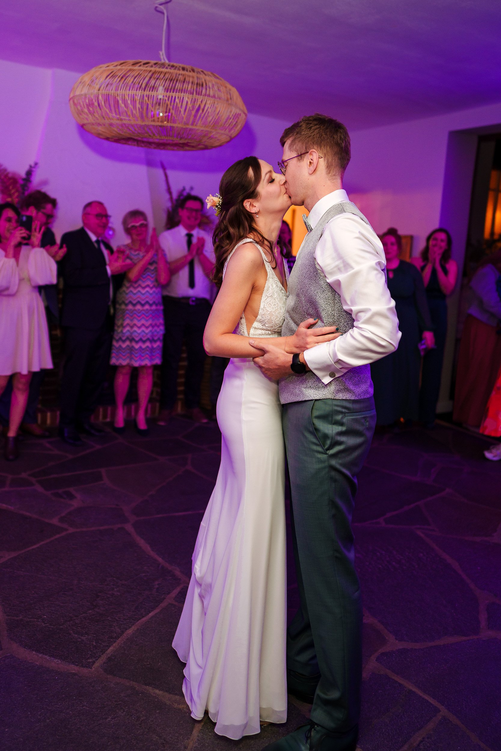 Ein Brautpaar tanzt bei einer Hochzeit, umgeben von Gästen, mit warmem Licht und einem großen runden Lampenschirm aus Holz im Hintergrund.