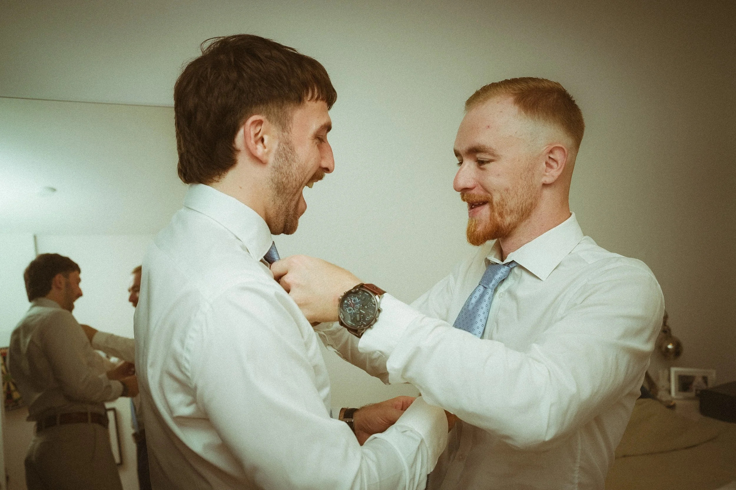 Zwei Männer in weißem Hemd und Krawatte lachen und umarmen sich, während einer dem anderen die Krawatte bindet.