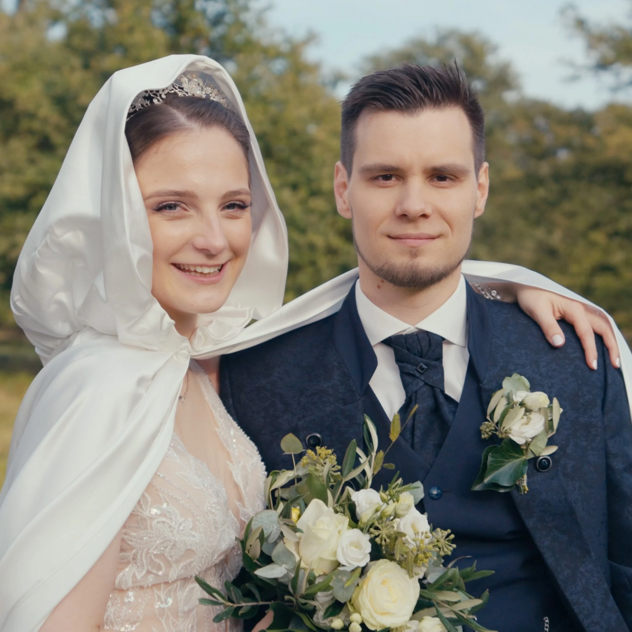 Ein Brautpaar bei ihrer Hochzeit, draußen im Grünen, der Braut trägt ein weißes Kleid mit Schleier und einen Umhang, der Bräutigam trägt einen dunklen Anzug mit Krawatte, sie hält einen Blumenstrauß, beide lächeln.
