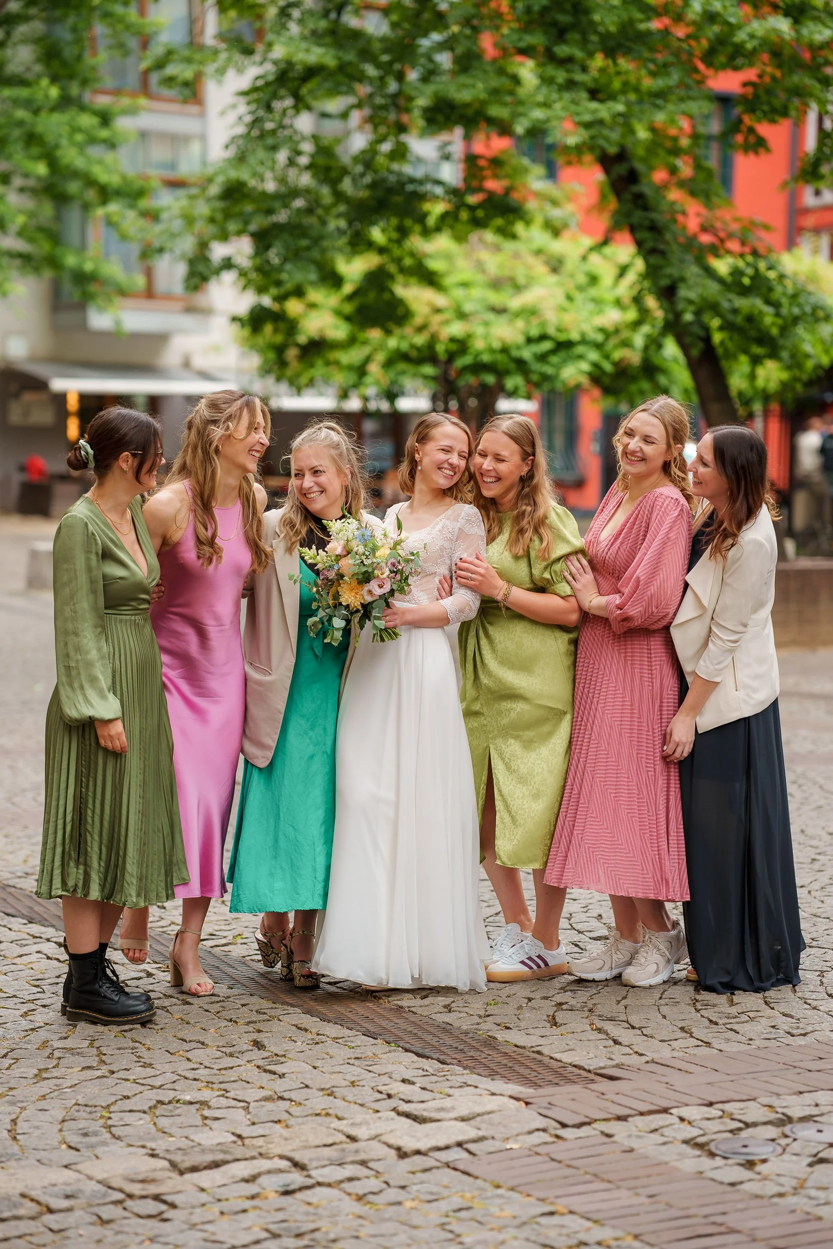 Eine Gruppe von acht Frauen, inklusive einer Braut, die in einem urbanen Außenbereich stehen und lachen. Die Braut hält einen Blumenstrauß, alle tragen farbenfrohe Kleider, und es gibt Bäume im Hintergrund.