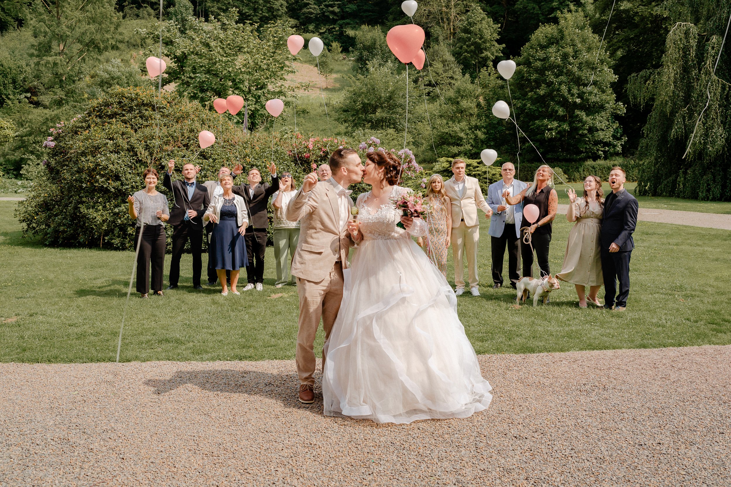 Brautpaar küsst sich vor Hochzeitsgesellschaft im Park, Heliumballons überall, sonniger Tag, grüne Bäume im Hintergrund.